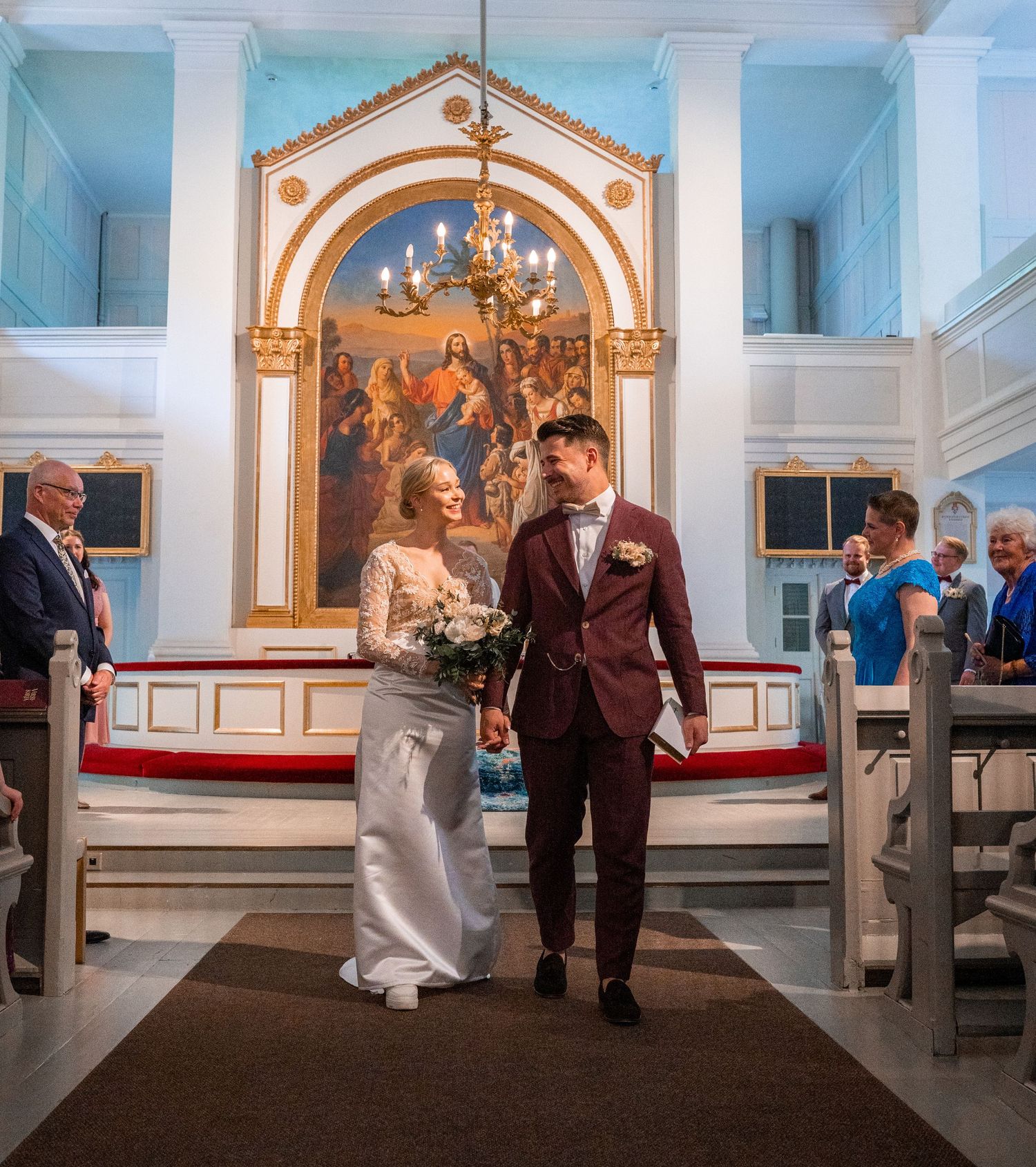 Bride and groom walk down church aisle after wedding ceremony with ornate altar and chandelier in background.