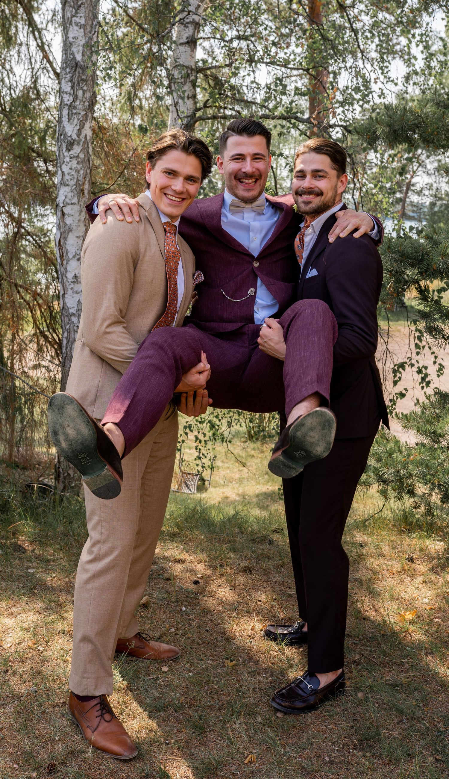 Two groomsmen playfully lifting the groom in a burgundy suit outdoors at a wedding.