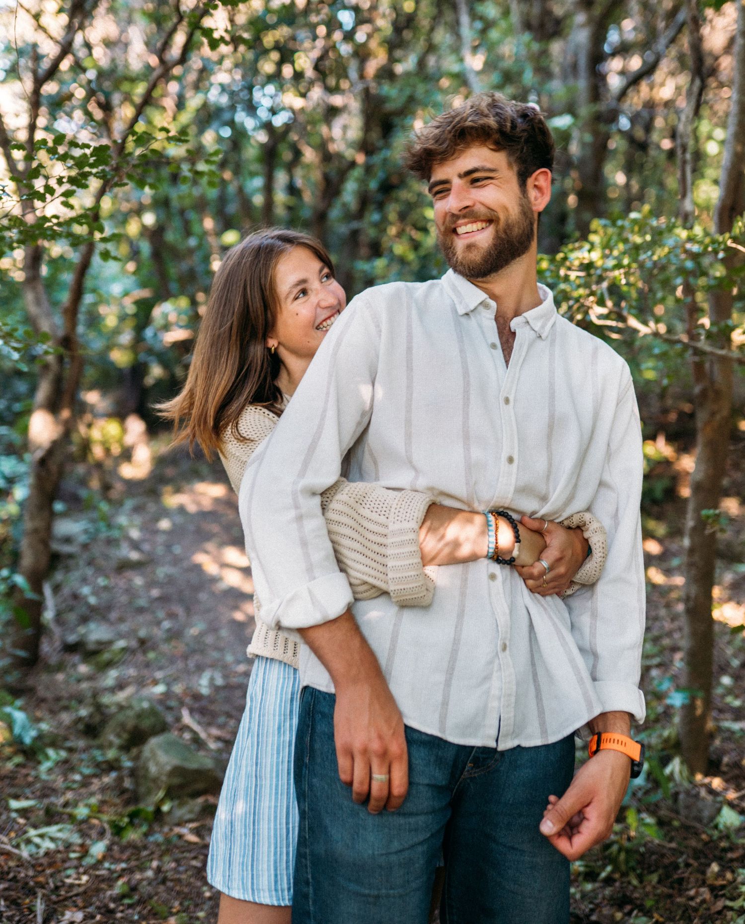 Happy couple embracing and smiling together on a sunlit forest path surrounded by trees.