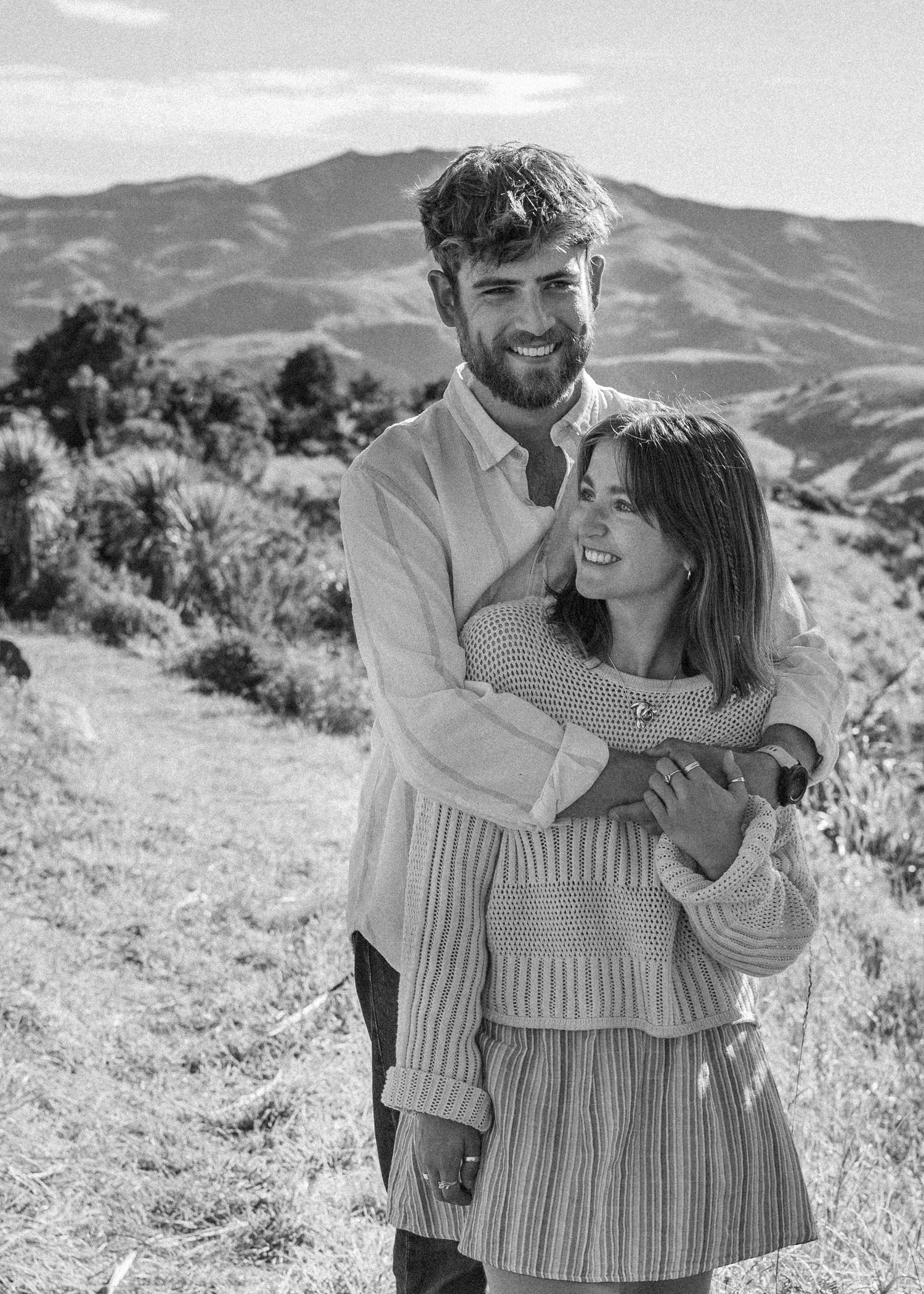 A smiling couple poses together in a black and white photo with scenic mountain hills in the background.