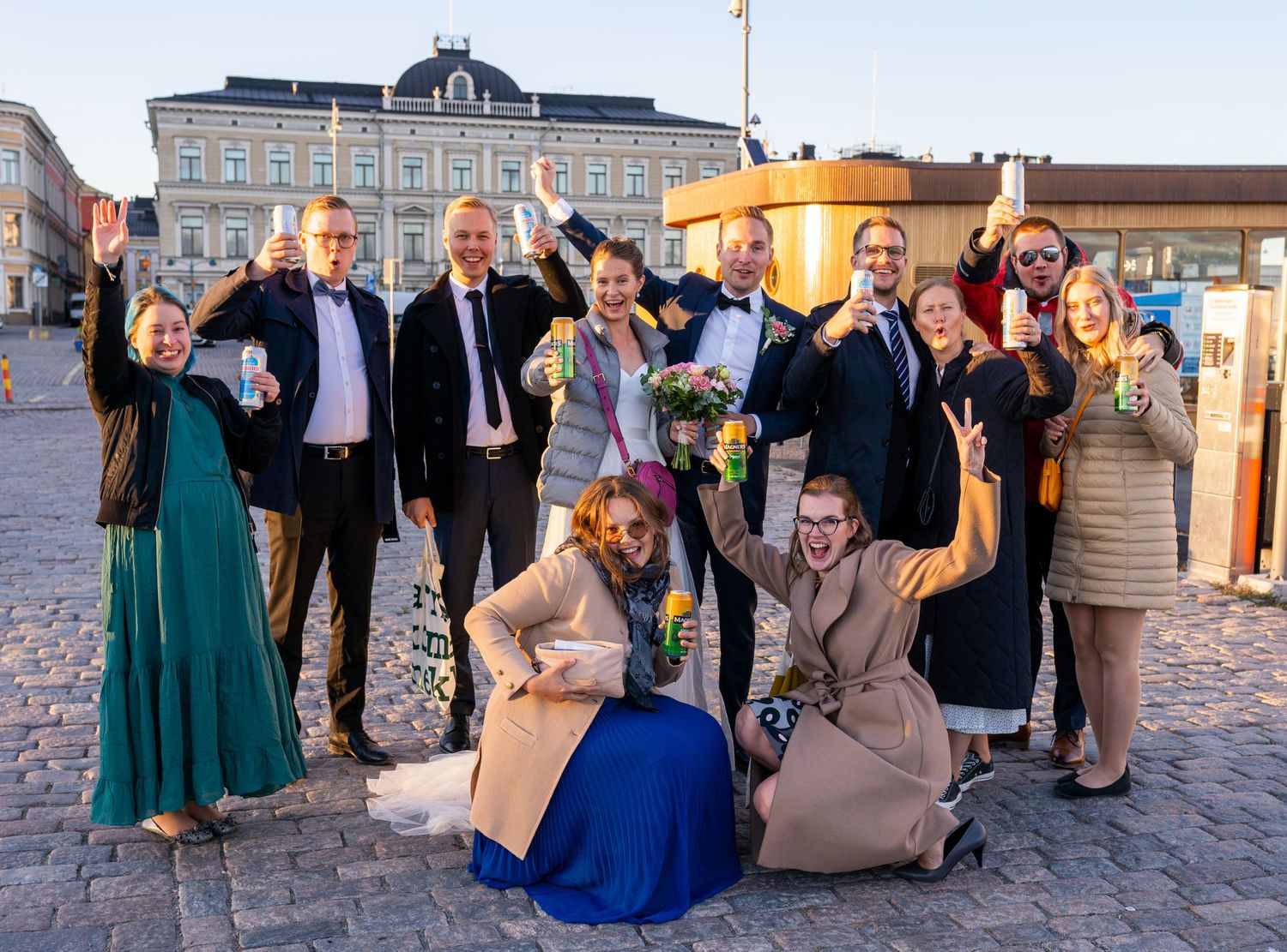 Wedding party celebrating outdoors in Helsinki city square, raising glasses and bouquets in joyful group photo.