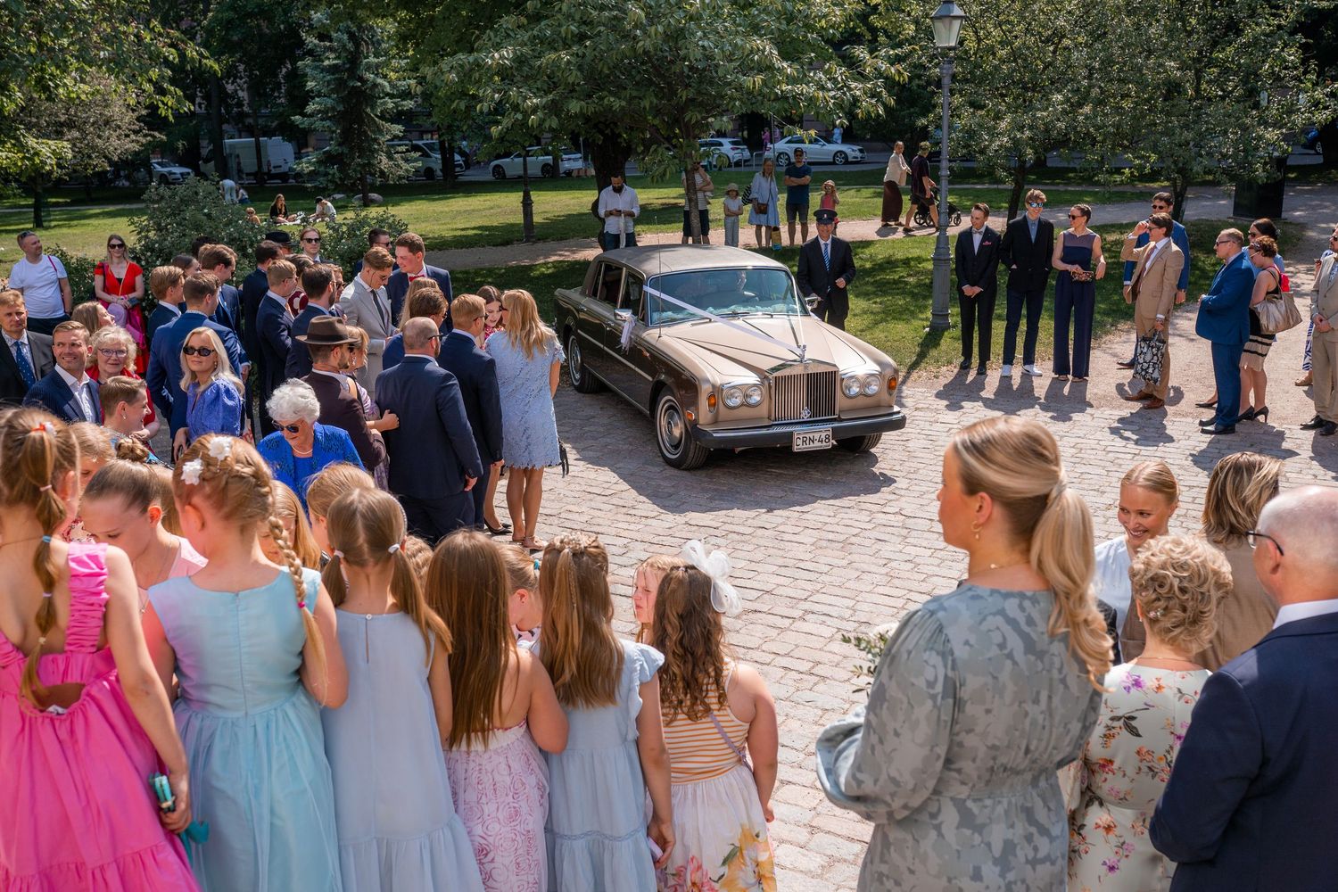 Vintage black wedding car arrives as guests gather on cobblestone path surrounded by lush trees.