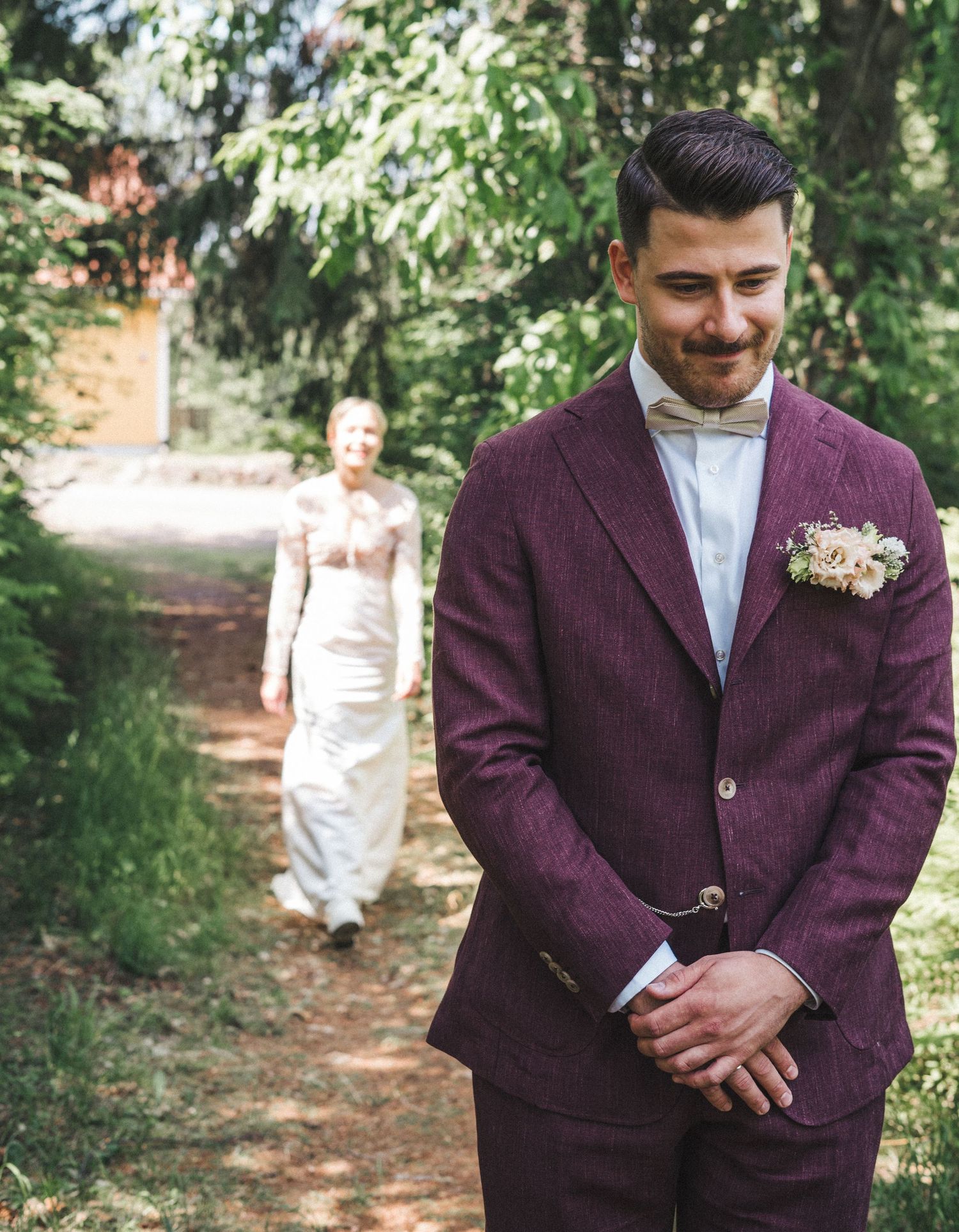 Groom in purple tweed suit with boutonniere awaiting bride approaching on a forest path.