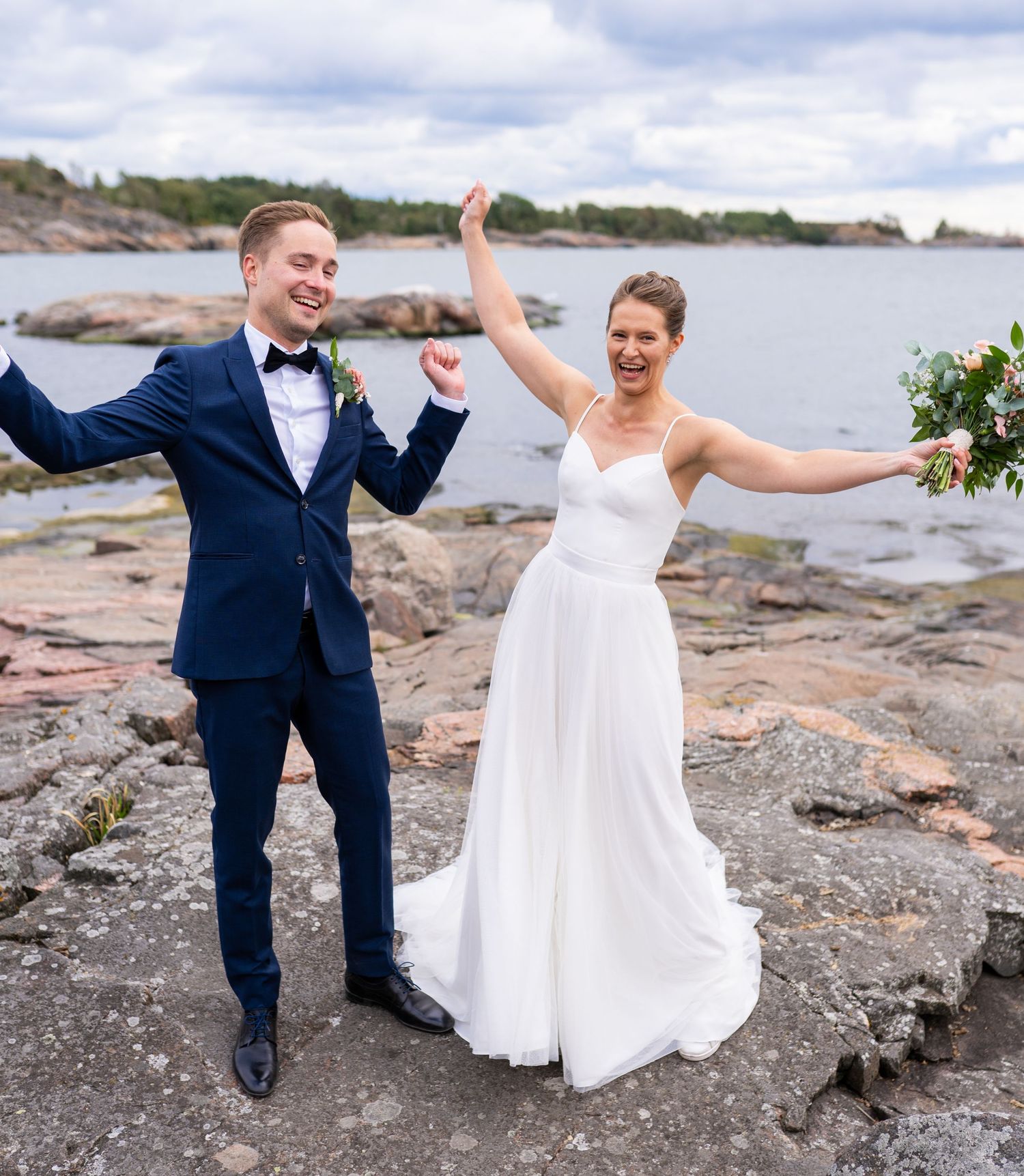 Joyful bride and groom celebrating on rocky seaside, arms raised, bride in white gown holding colorful bouquet.