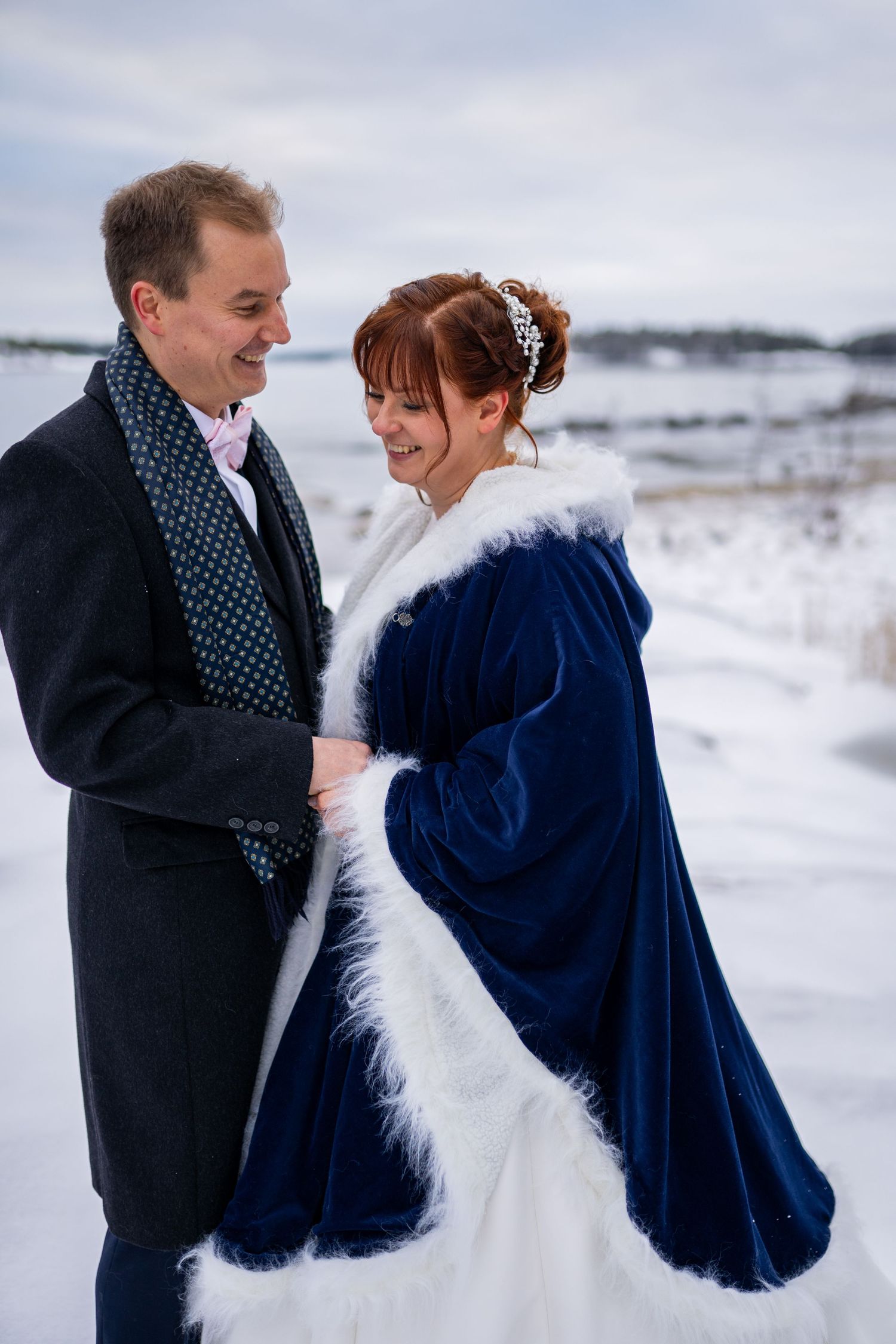 Smiling bride in navy blue fur-trimmed cape and groom in gray suit laugh together in a snowy winter landscape.