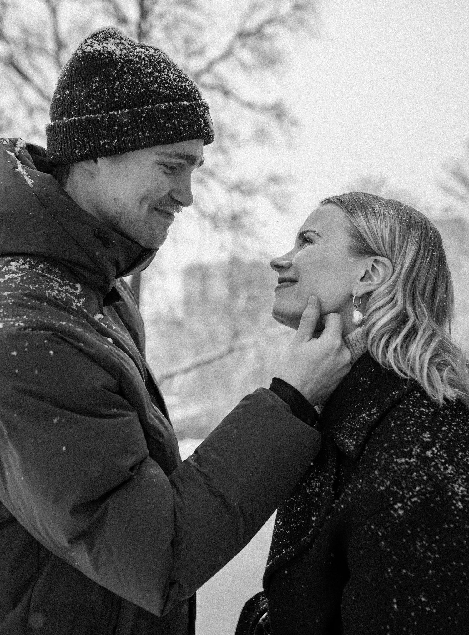Black and white winter photo of smiling couple, woman tenderly touching mans face in snowy outdoor setting.