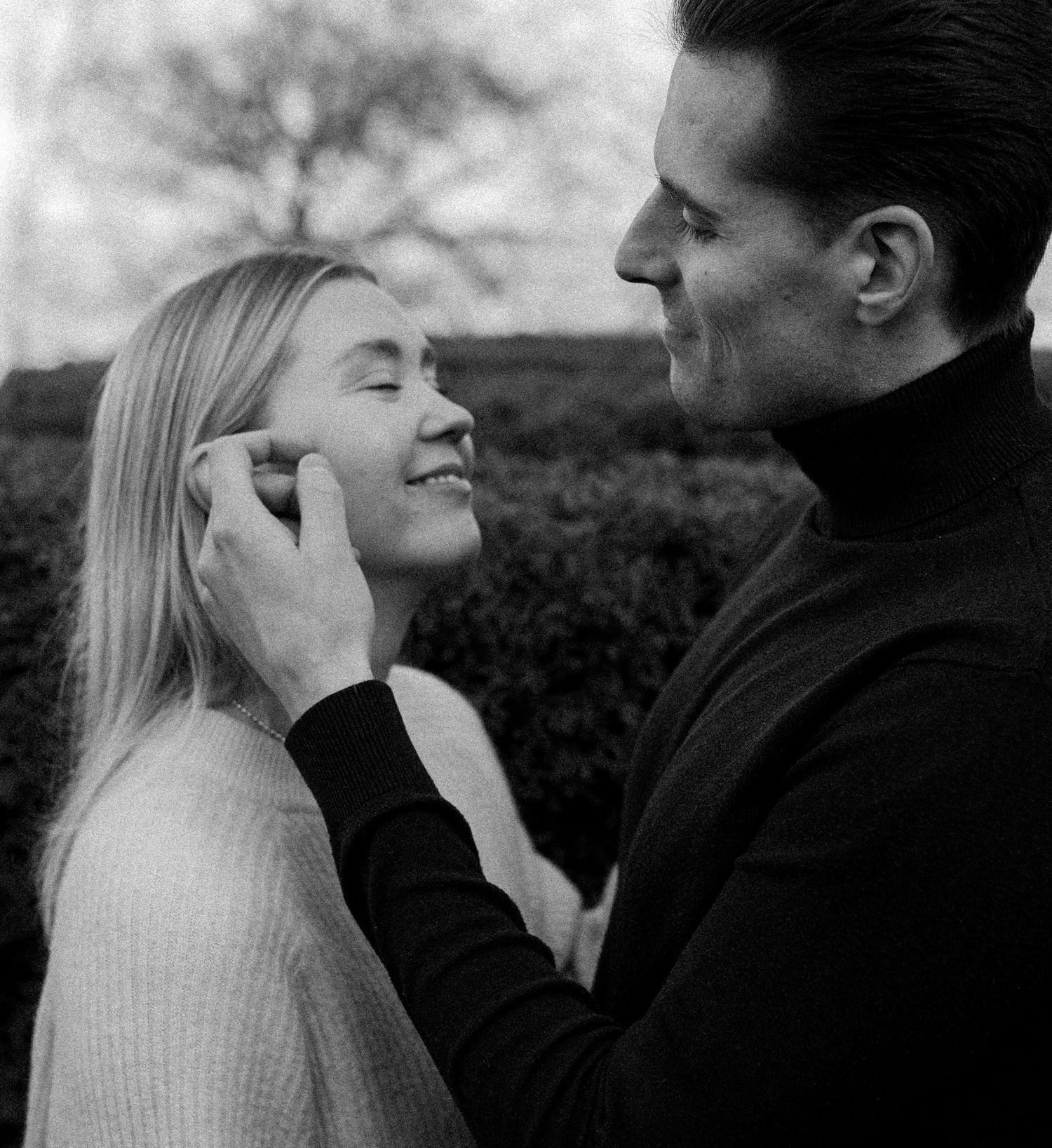 Black and white photo of a smiling blonde woman and a man in a turtleneck tenderly touching her face outdoors.