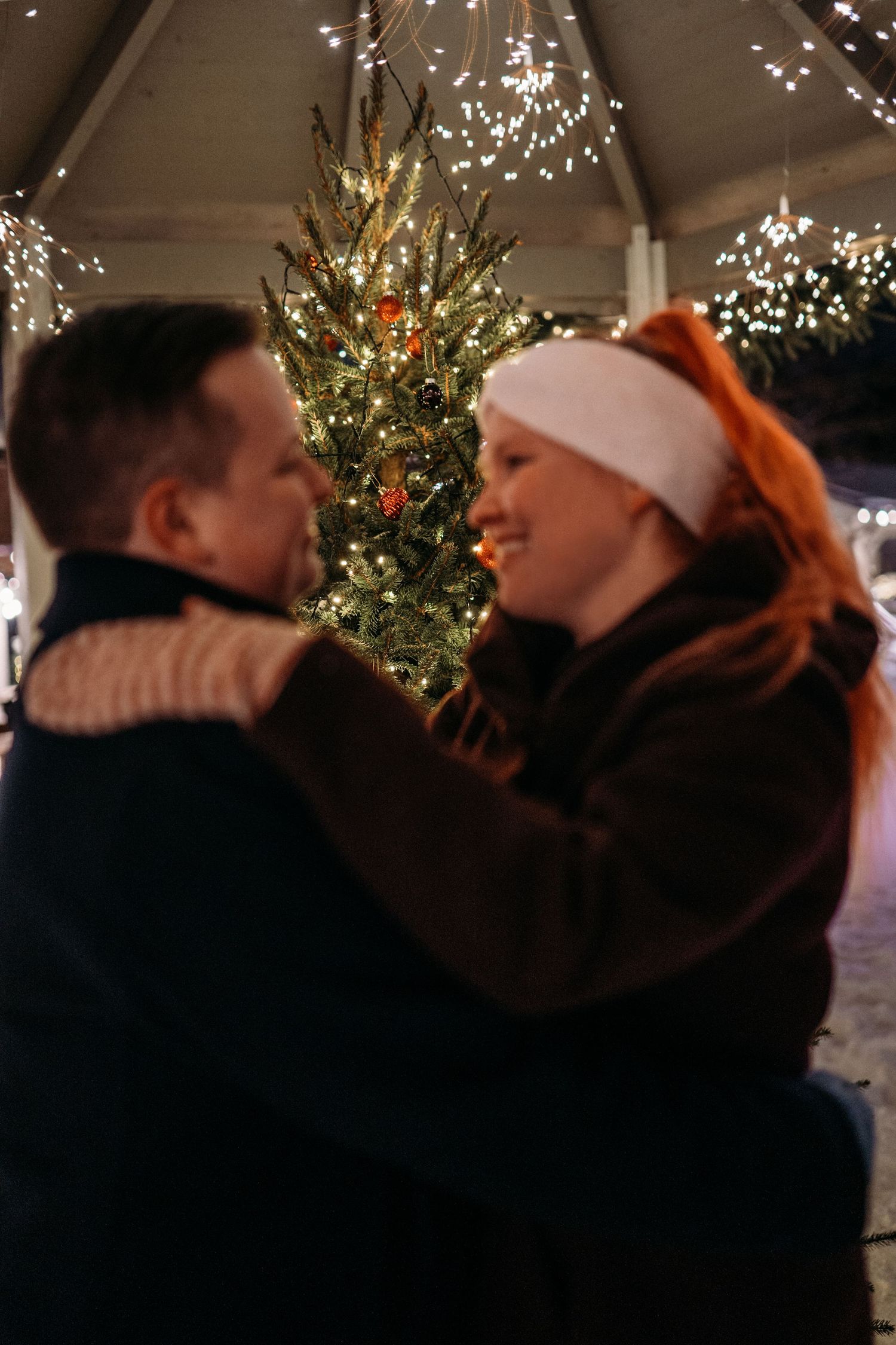 Couple embracing near a decorated Christmas tree with warm lights at a festive outdoor holiday market.
