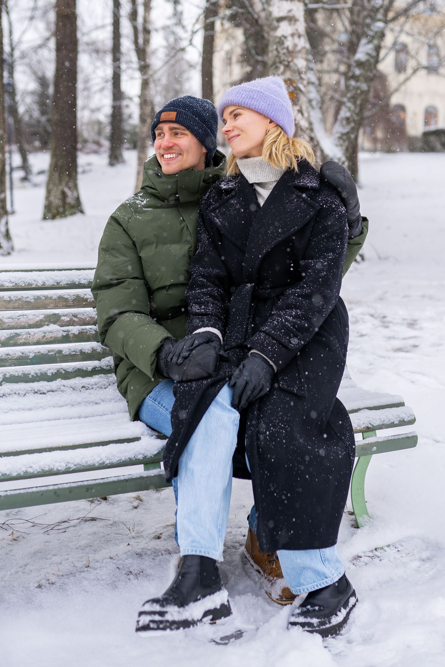 Couple sitting on a snowy park bench in winter, wearing warm coats and hats, smiling and holding hands.