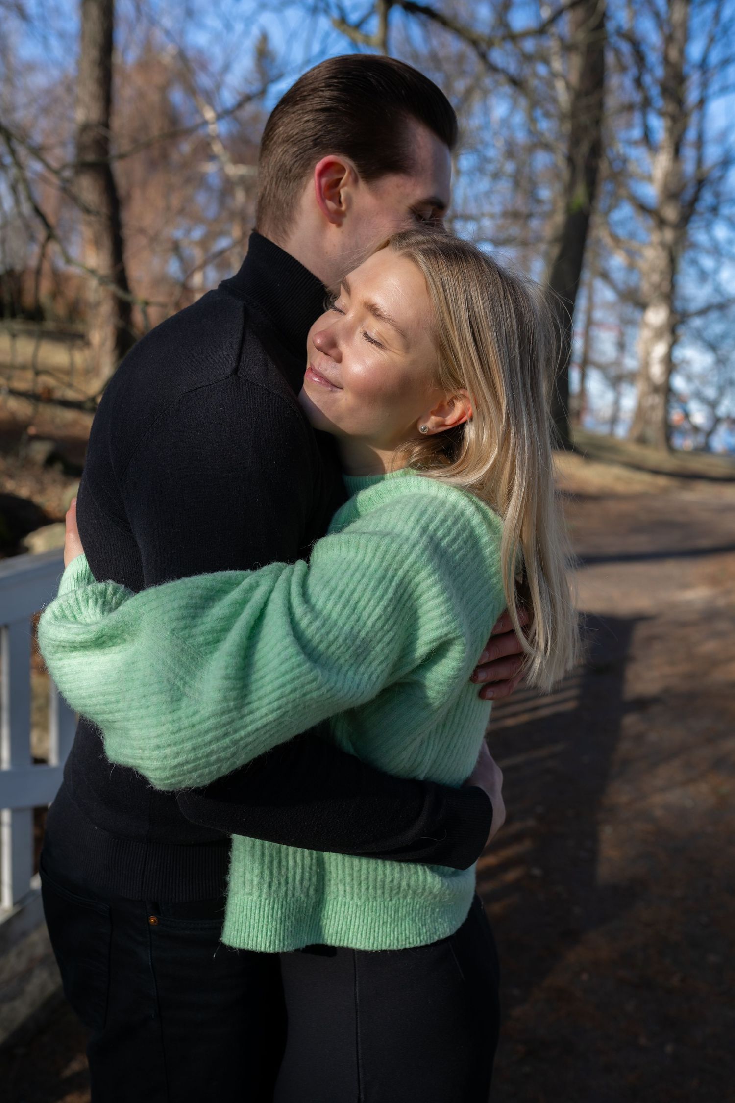 A couple embracing outdoors in a park, woman in mint green sweater smiling with eyes closed, man in black turtleneck.