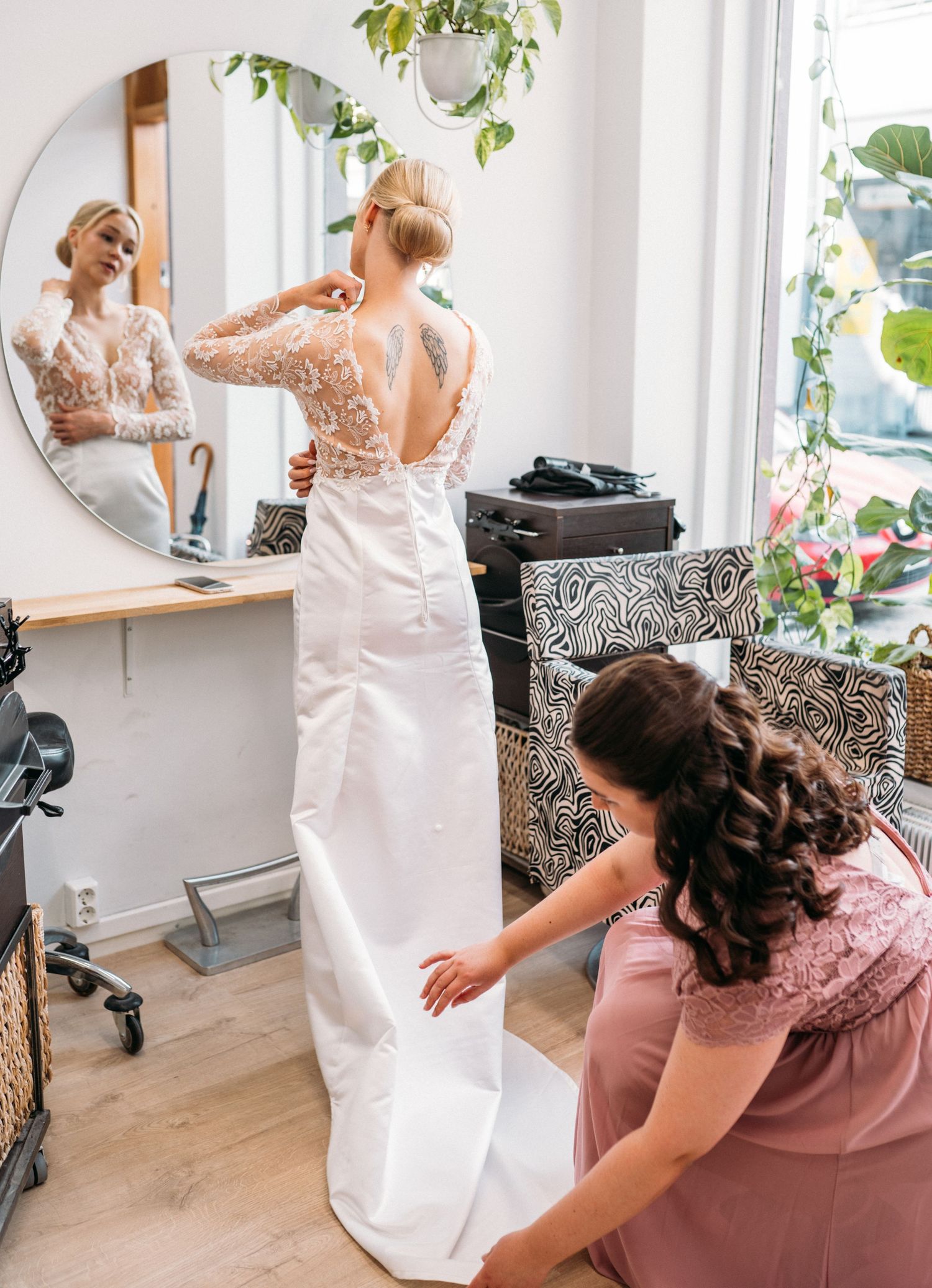 Bridesmaid in pink dress adjusting the train of a brides white lace wedding gown by a mirror.