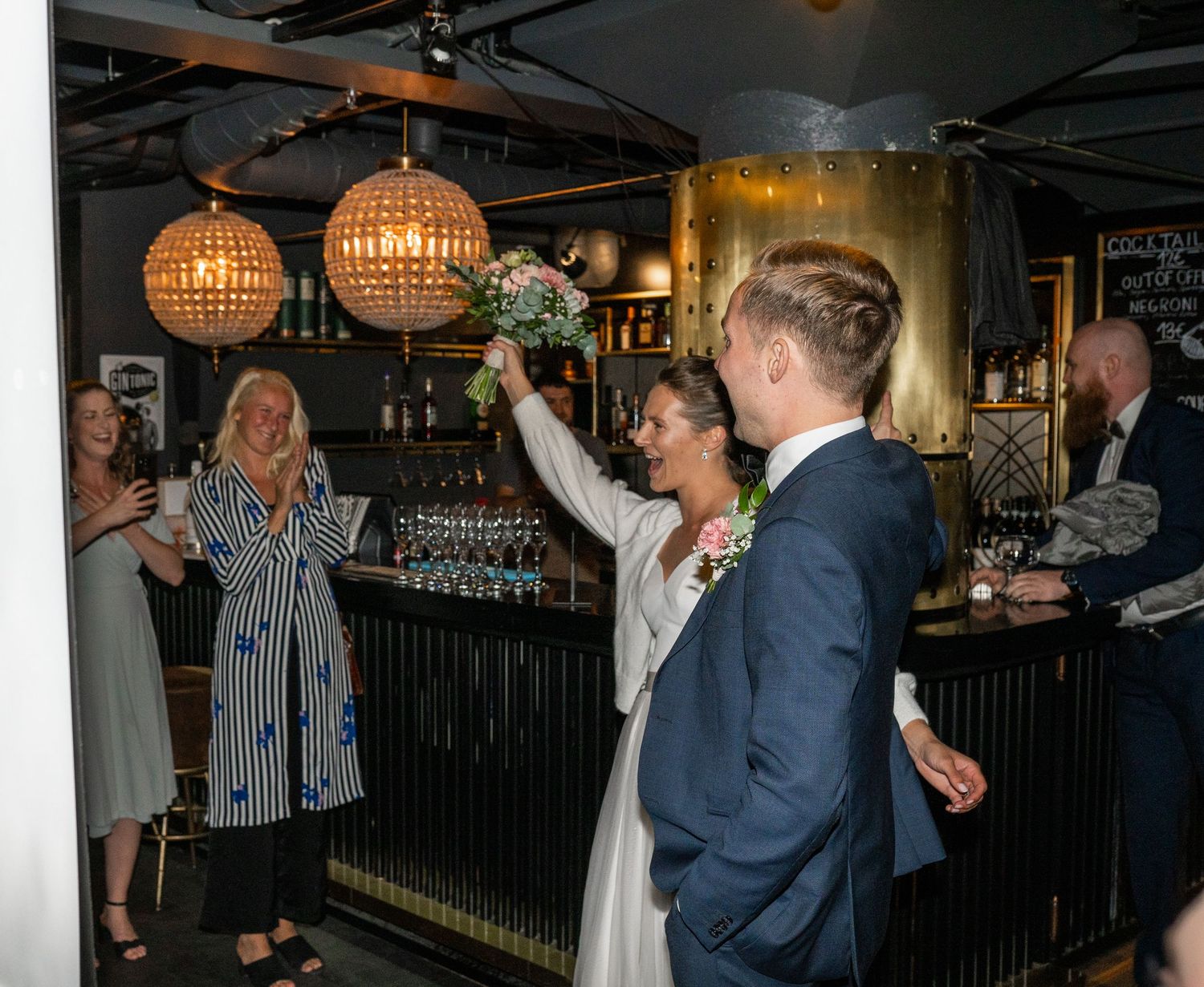 Bride raises bouquet triumphantly at bar while guests applaud the newlyweds at wedding celebration.