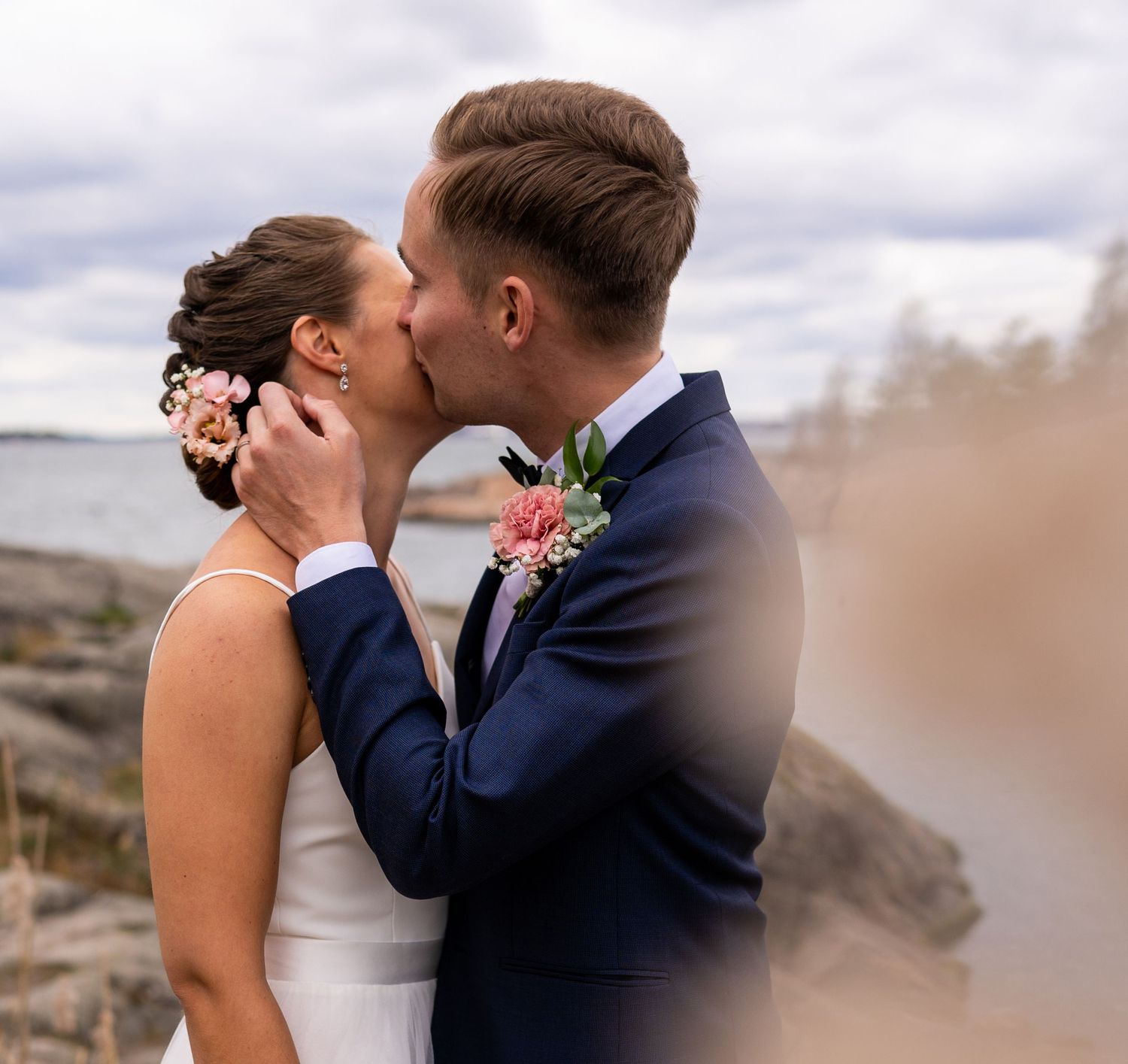 Bride and groom sharing a romantic kiss outdoors near rocky shoreline, both dressed in wedding attire.