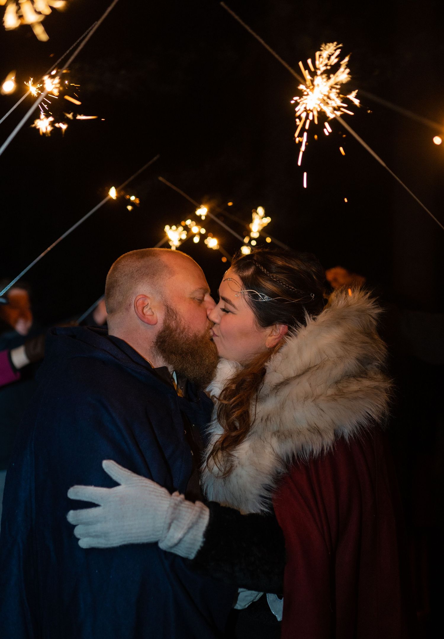 Couple kissing surrounded by sparkling sparklers at night, woman in fur-trimmed coat and white gloves embracing bearded man.