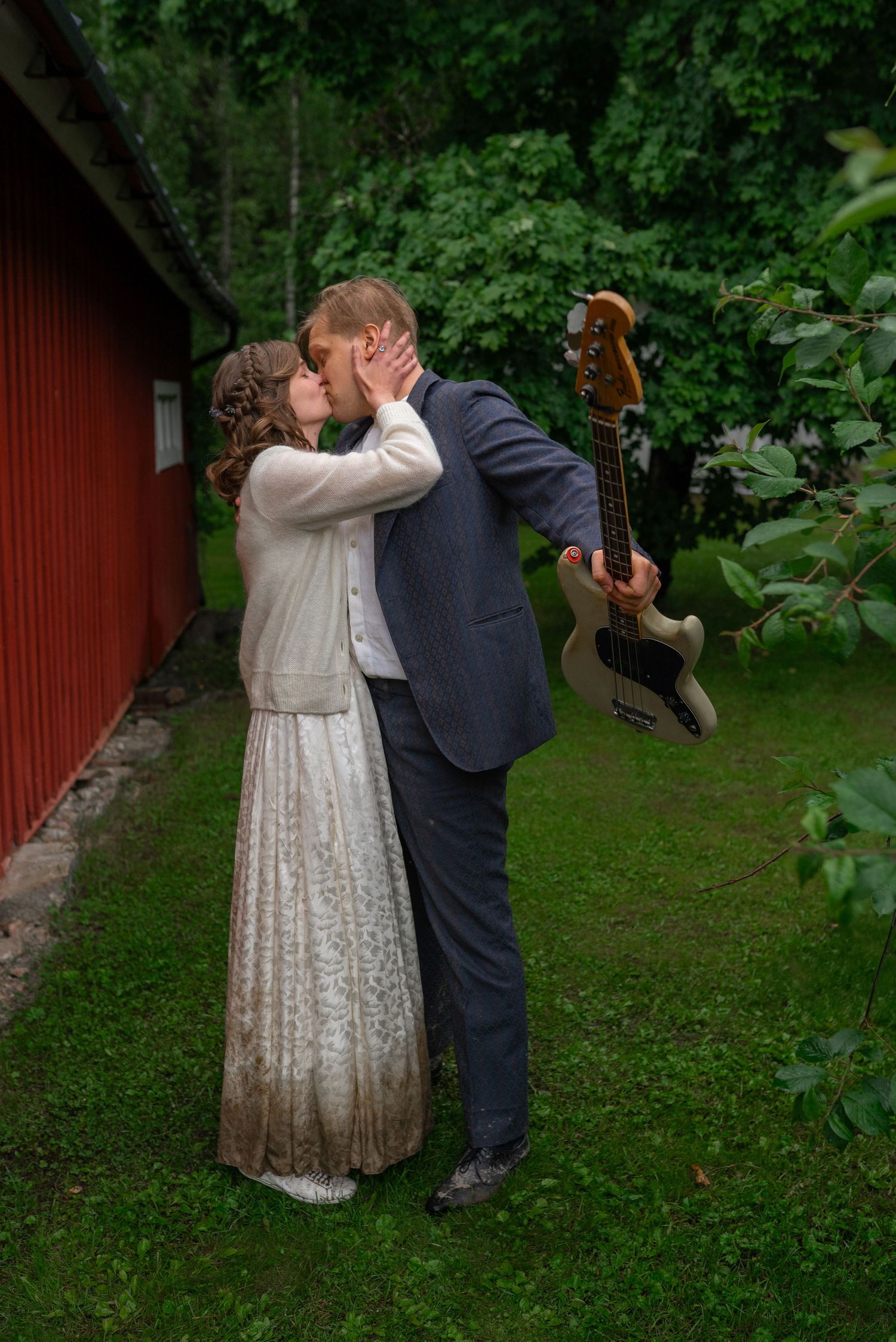 Groom holding a guitar shares a tender kiss with his bride on a lush green lawn.
