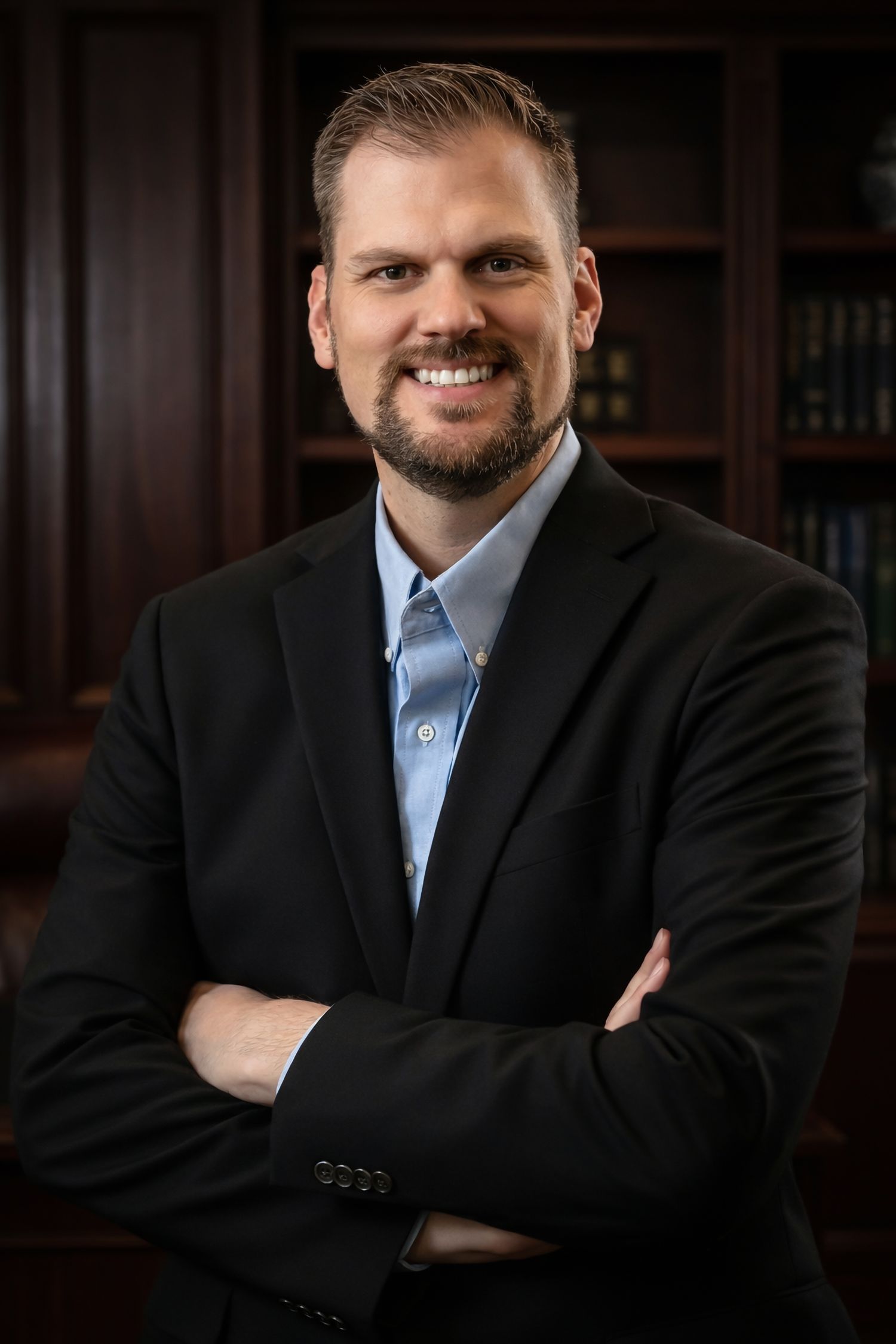 Professional headshot portrait of person in dark suit and blue shirt against wood paneled background.