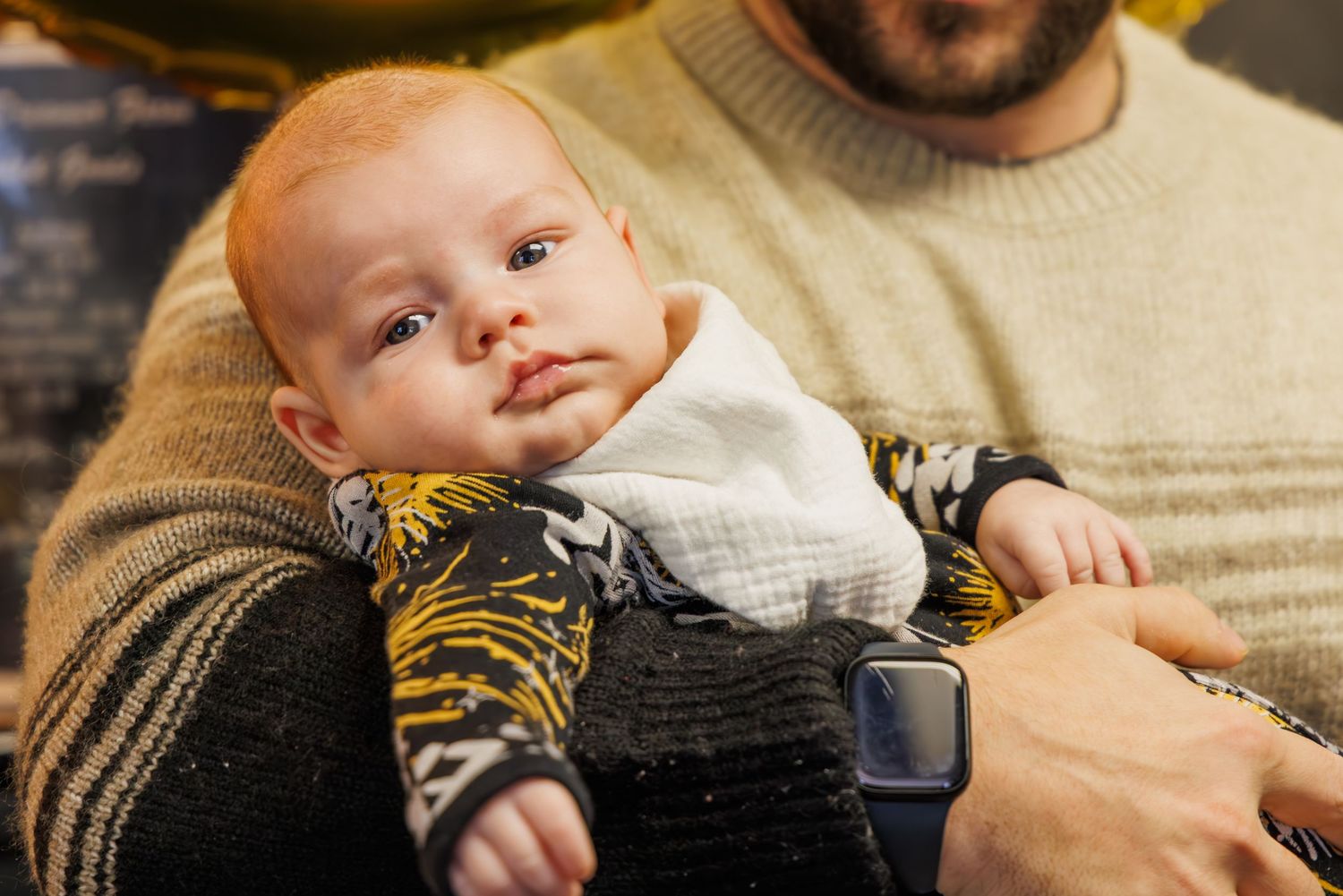 A baby in a striped onesie lies on a soft surface while being held.