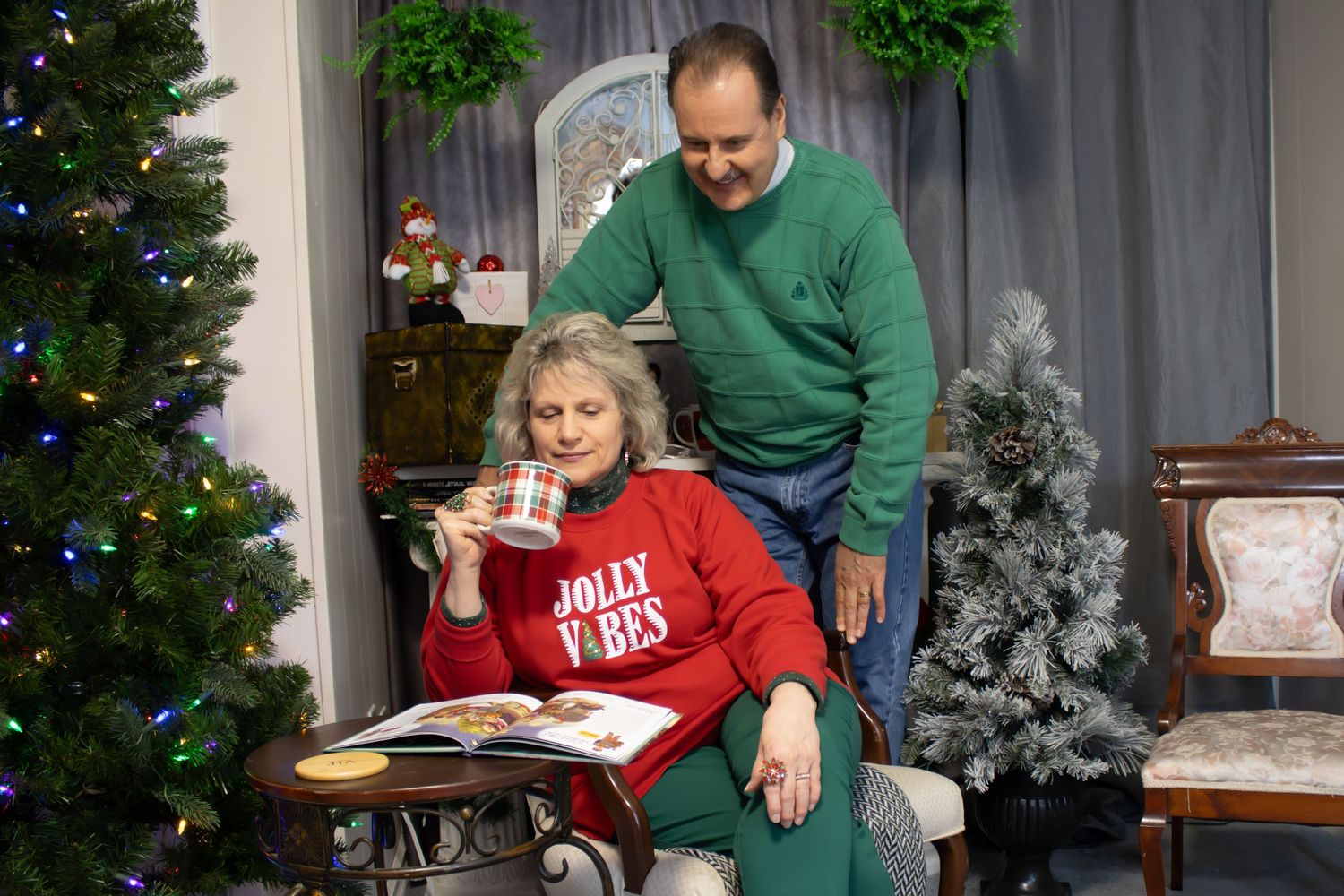 A cozy Christmas scene showing two people in holiday attire sitting and standing by decorated trees.