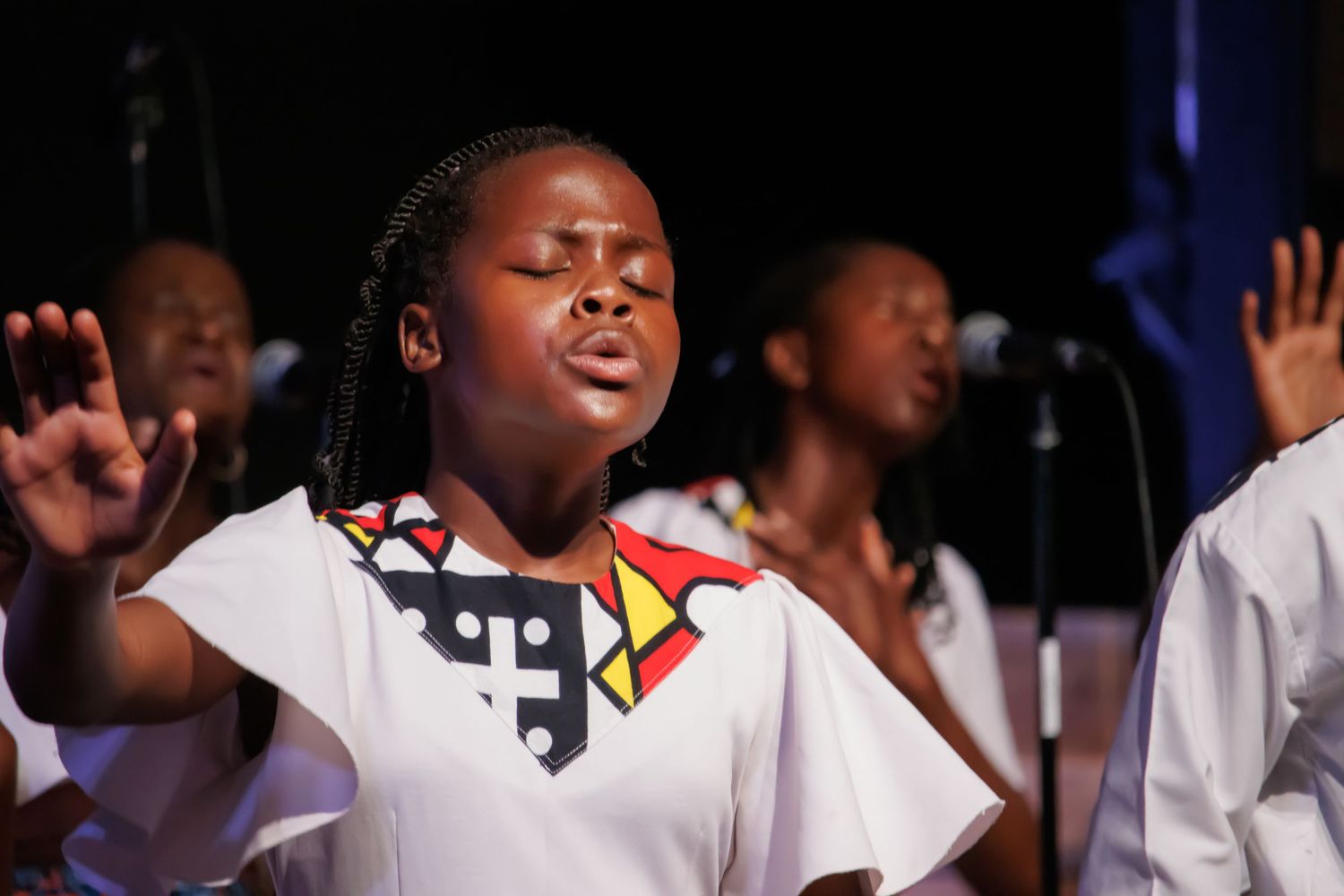 Gospel choir members in white attire singing with expressive passion on stage.