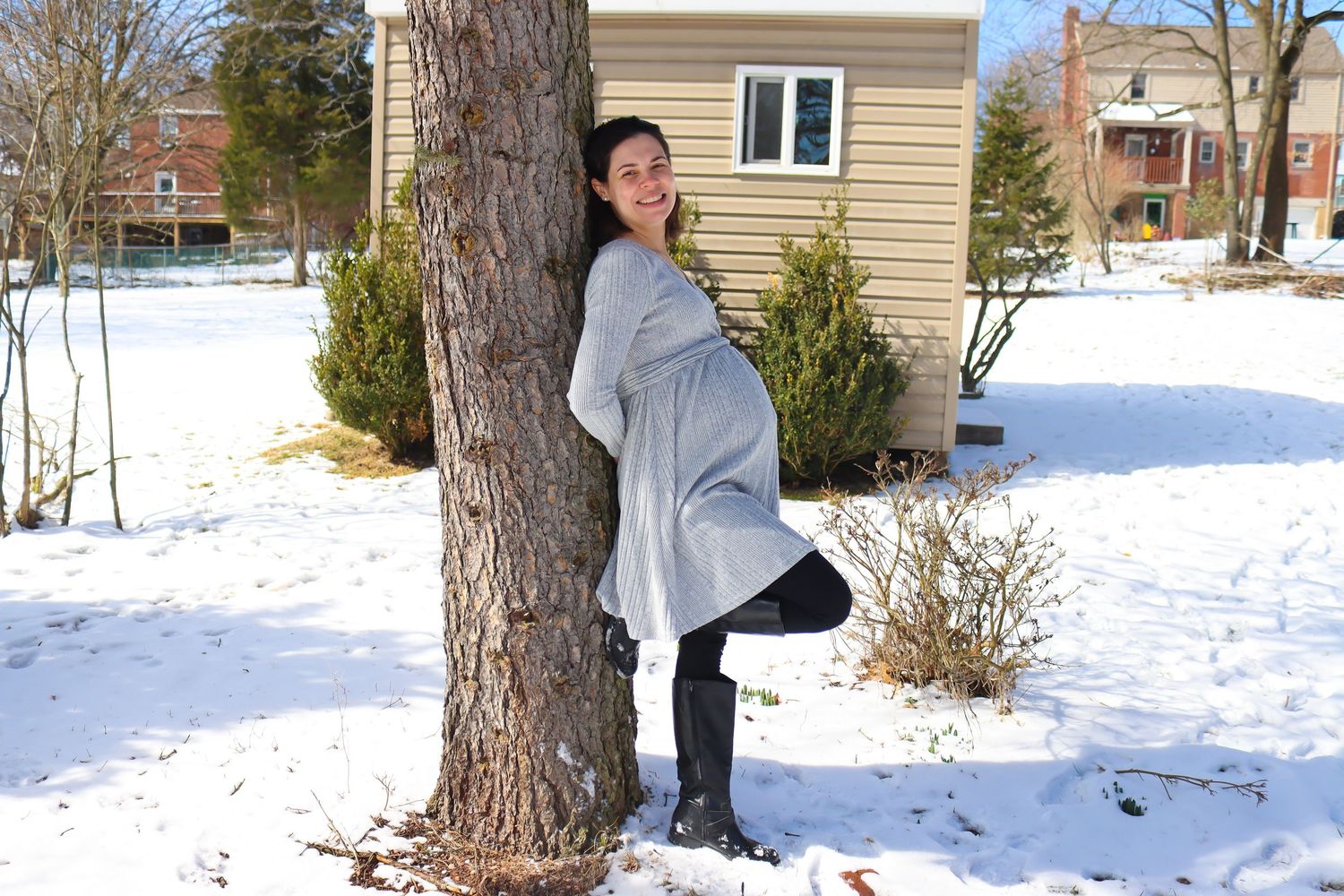 Person in gray clothing posing near a snow-covered house and pine tree.