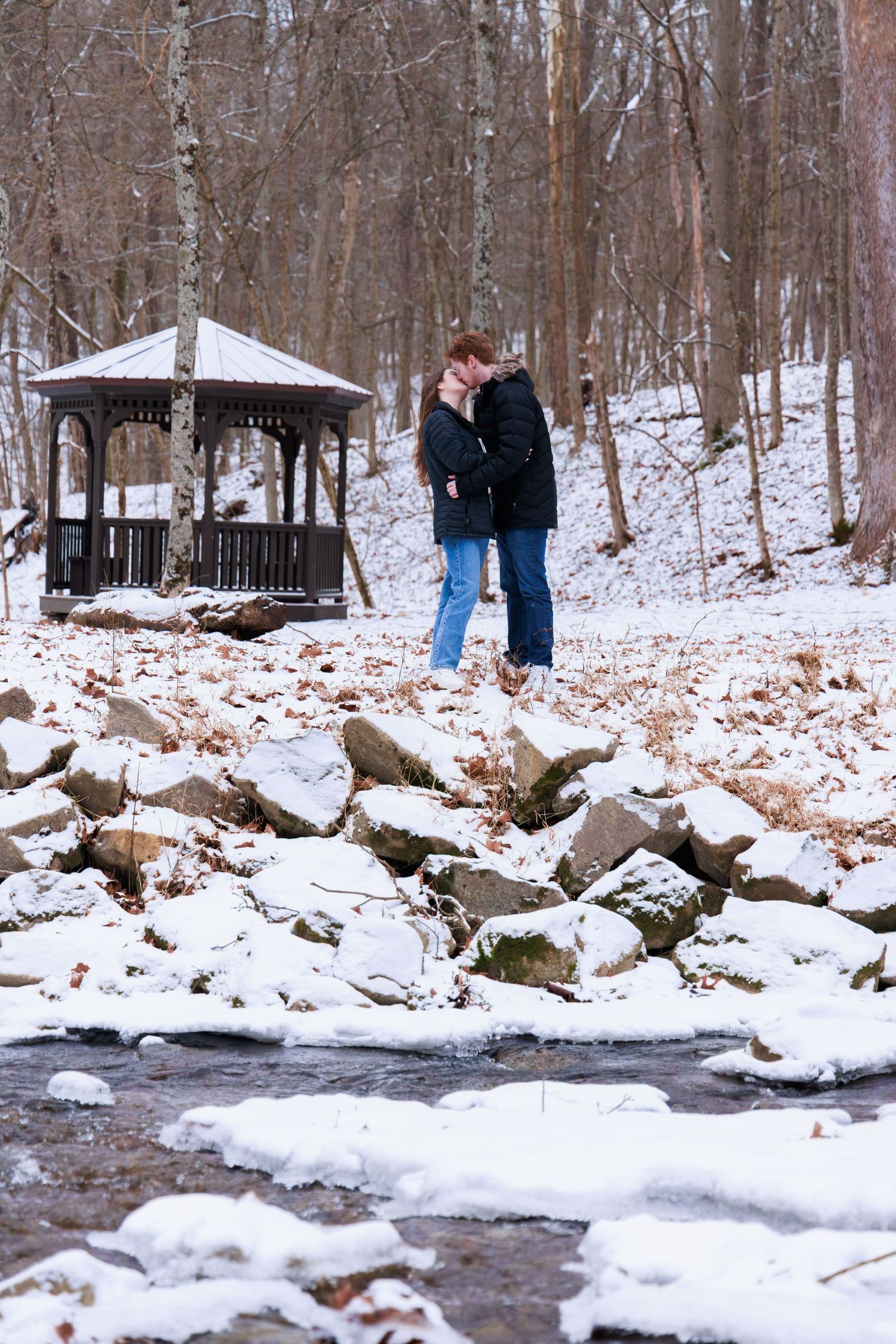 A couple stands near a rocky stream bank in a snow-covered forest with a gazebo in the background.