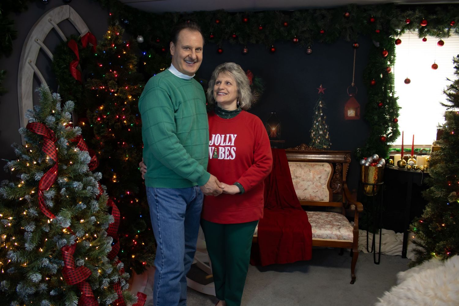 A festive holiday portrait with two people in red and green sweaters posed by decorated Christmas trees.