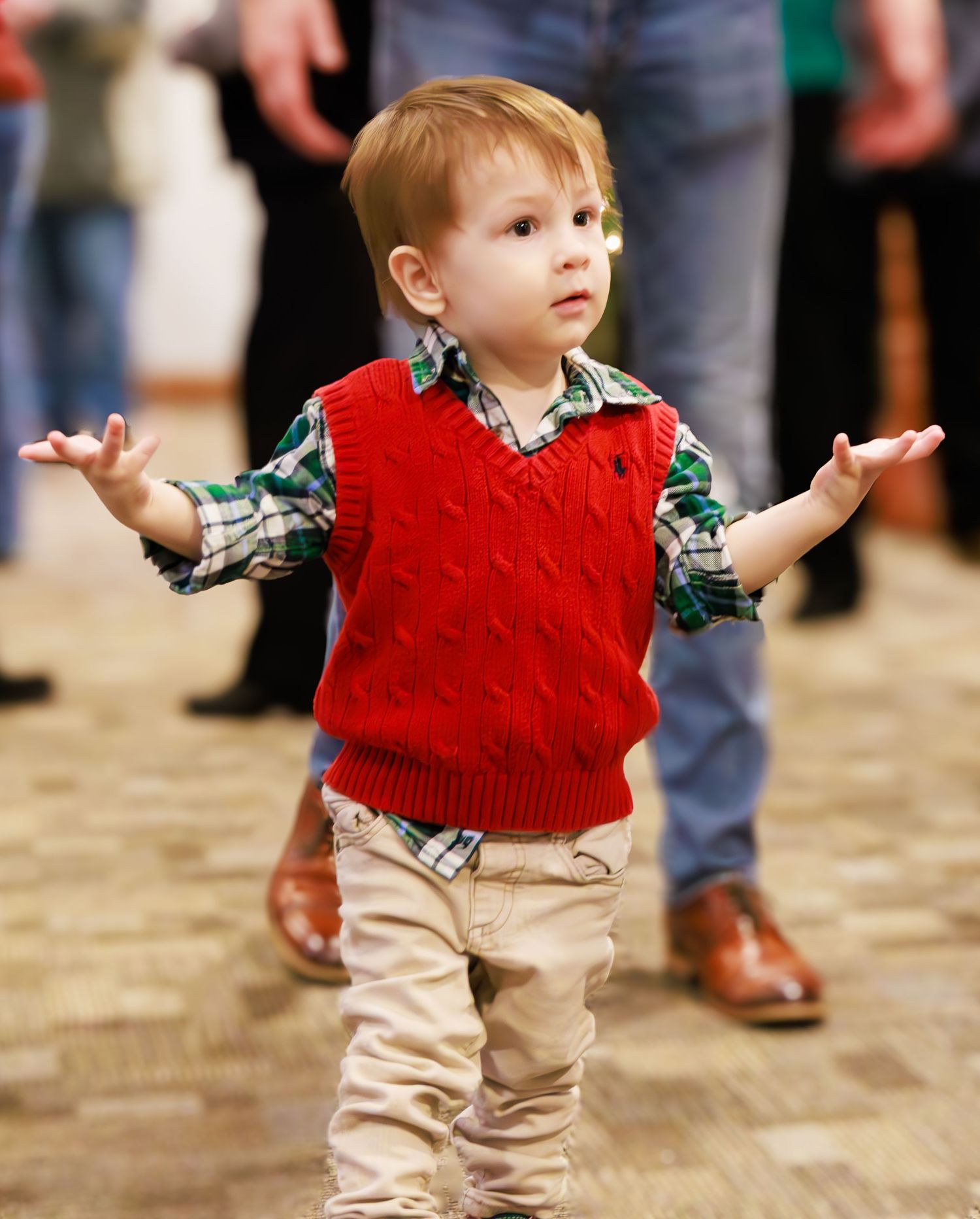 A toddler in a red vest and khaki pants walks across a carpeted floor.