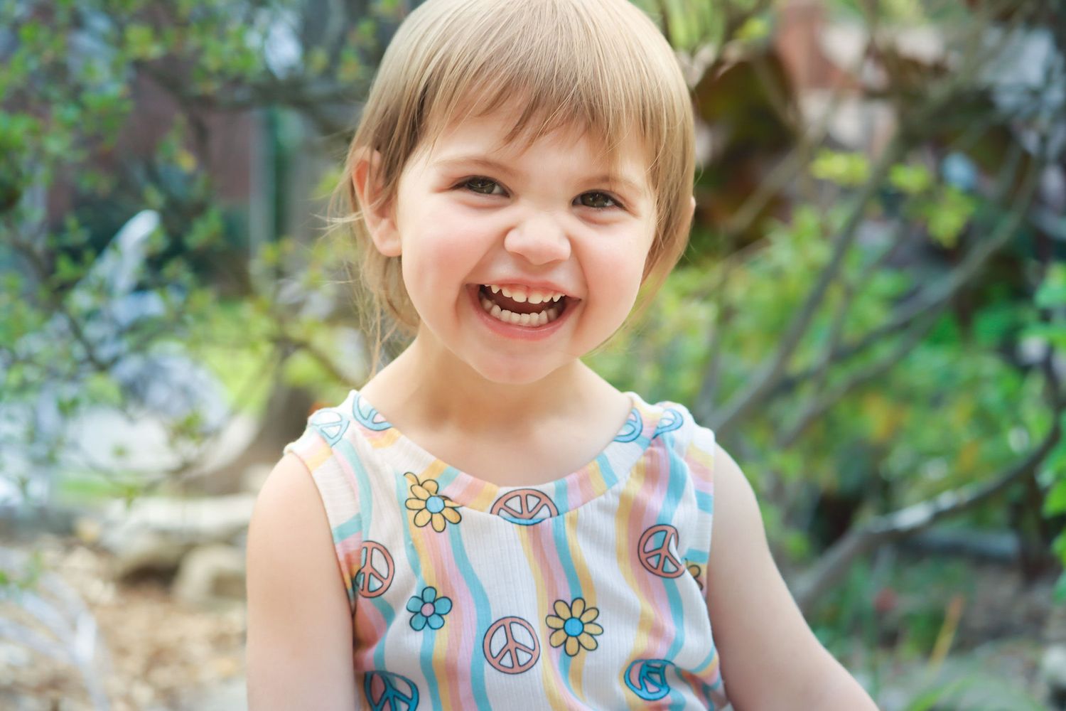 A child wearing a floral dress smiles joyfully while playing outdoors in natural lighting.