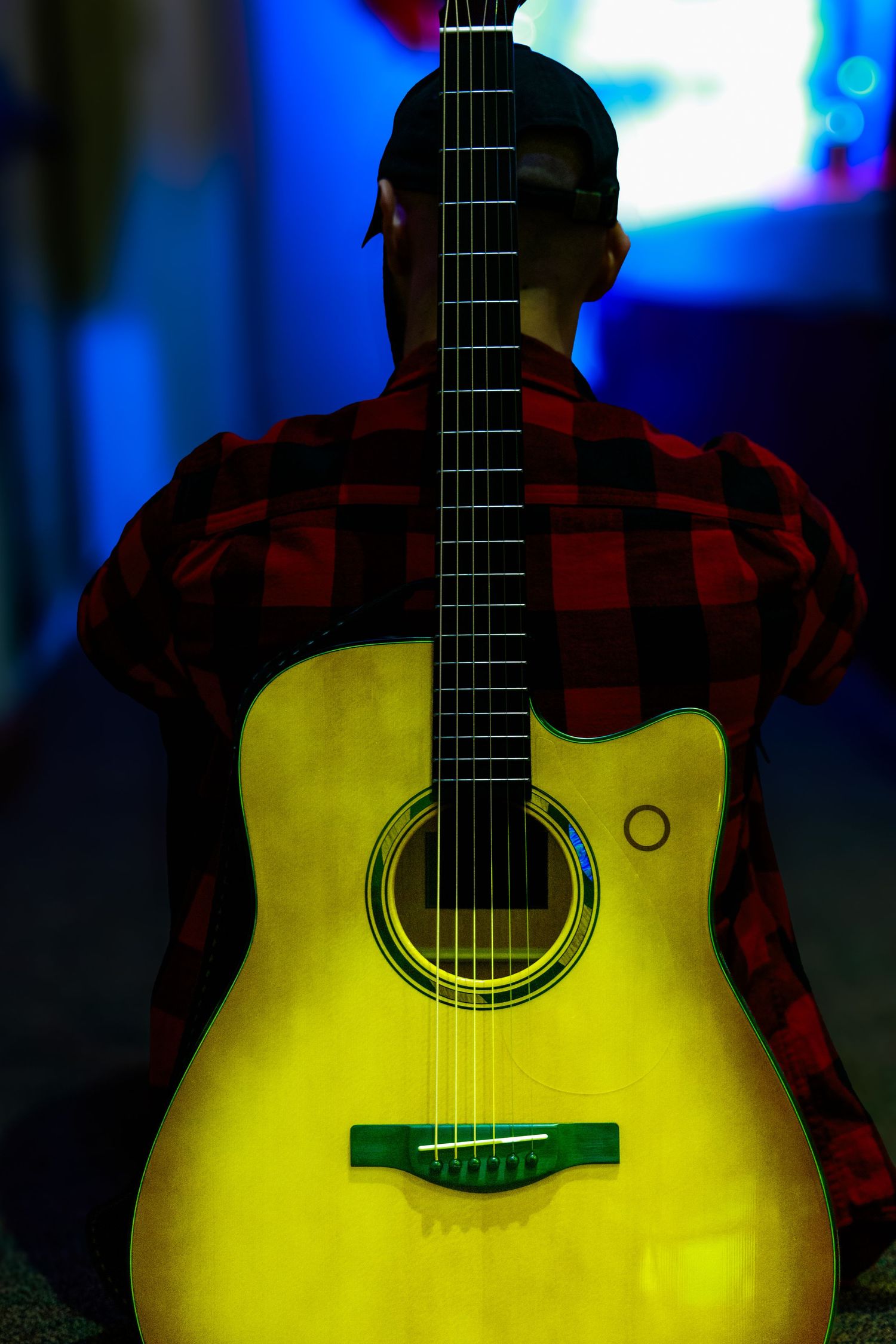 Yellow acoustic guitar glowing against dark background with performer in plaid shirt.