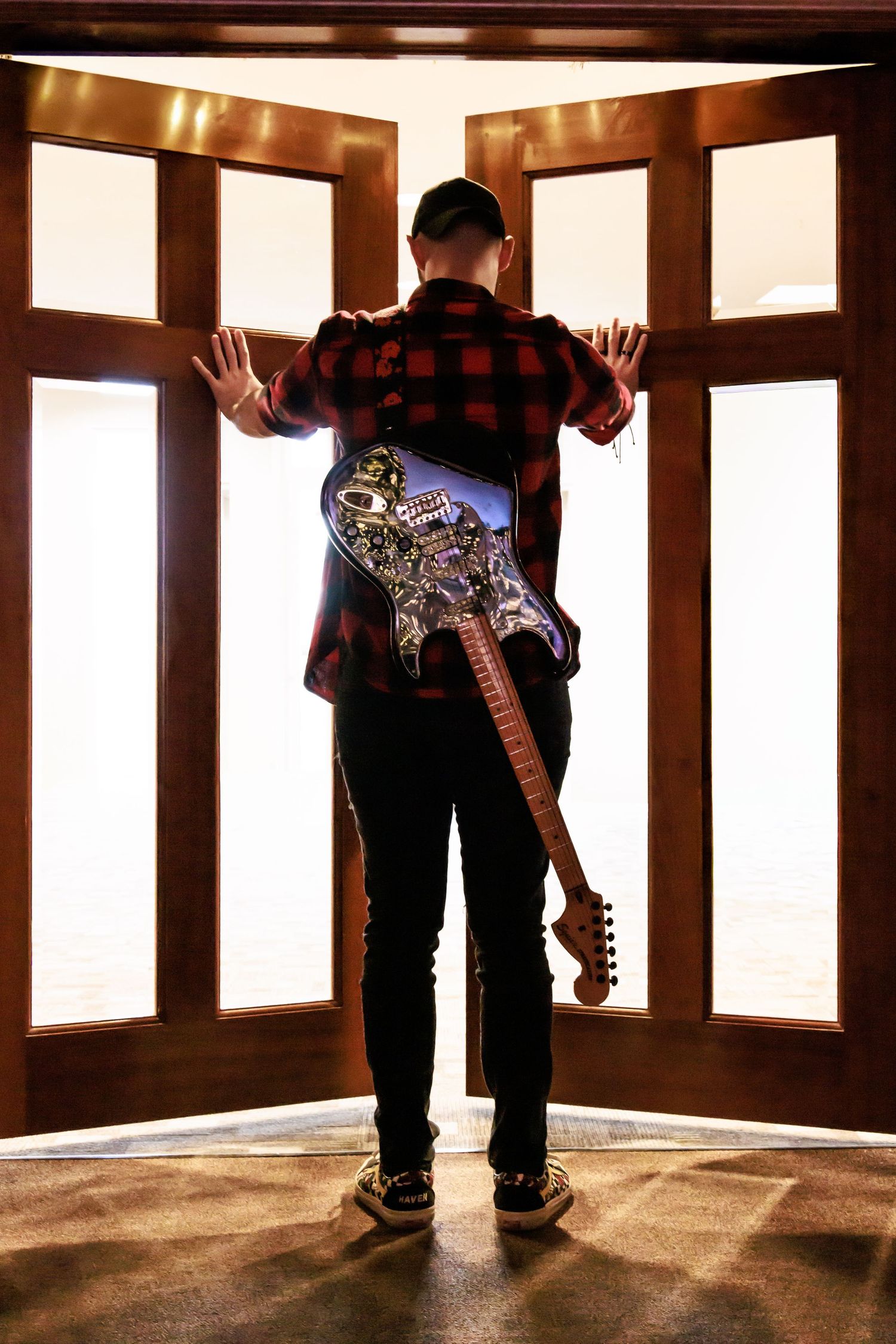 Musician wearing plaid shirt and dark jeans stands in front of illuminated windows with electric guitar strapped to back.