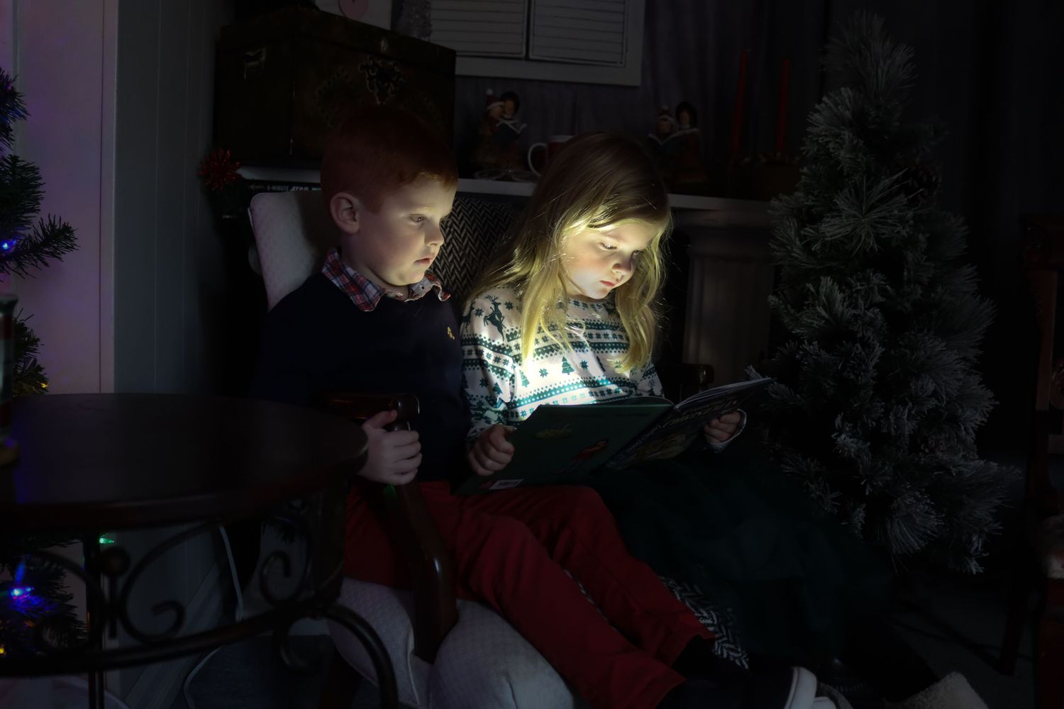 Two children share a tablet device in a dark room illuminated by Christmas tree lights.
