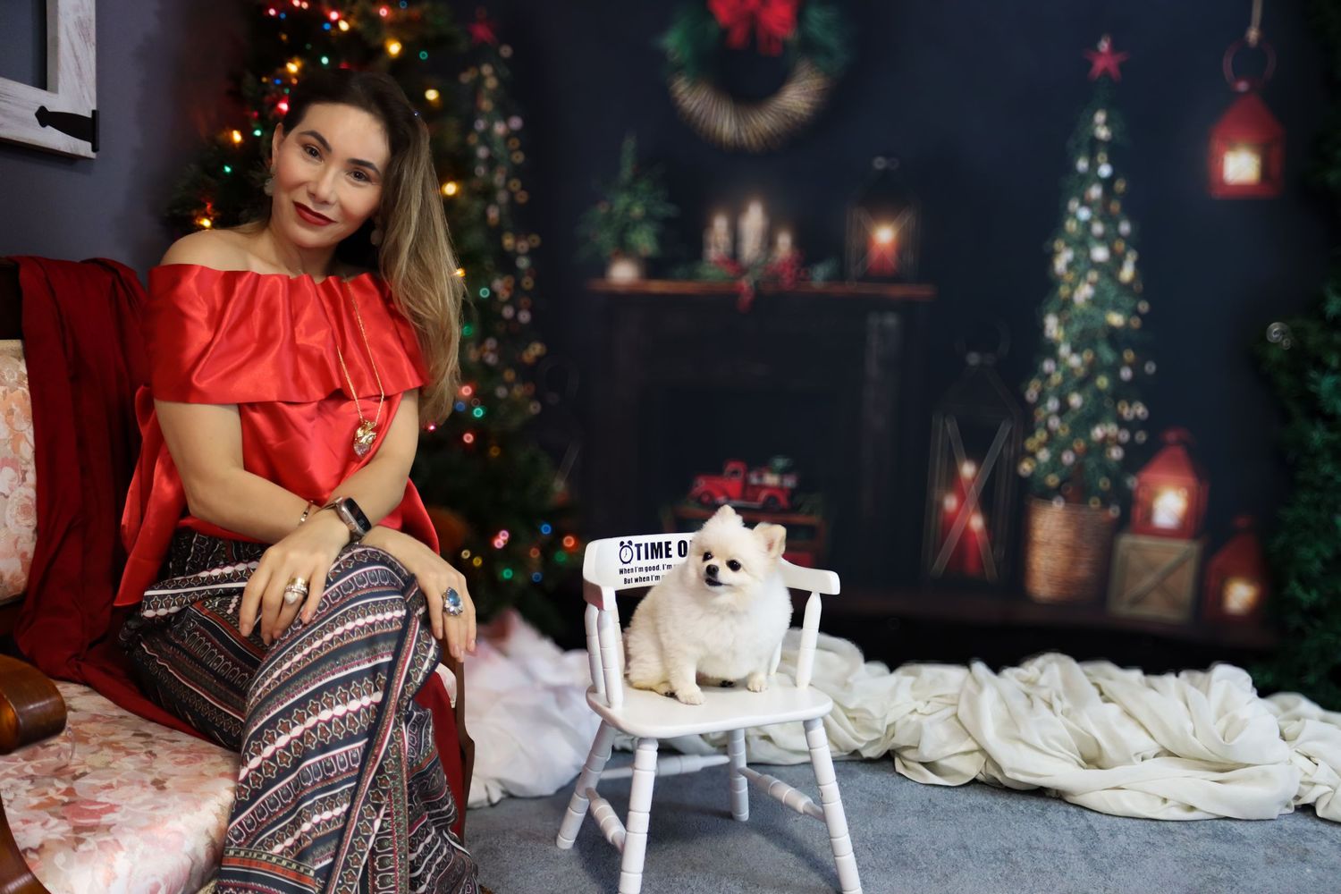 A holiday portrait featuring a woman in red beside a small white chair with a Pomeranian.