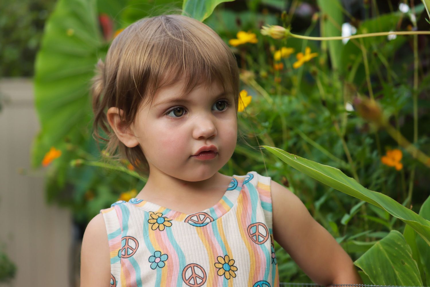 Child in peace sign print dress standing among garden flowers and greenery.