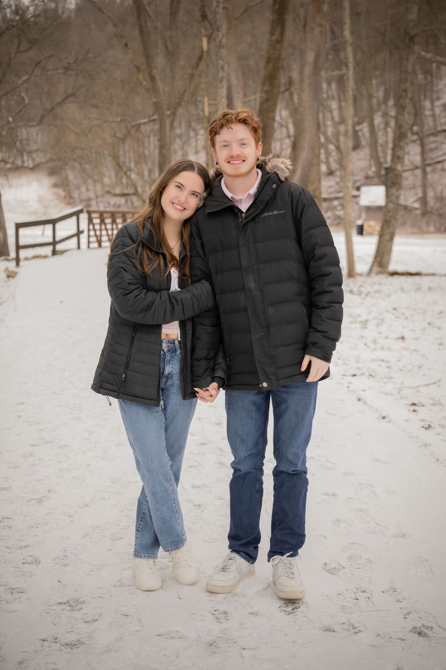 A couple in winter coats and jeans stands together on a snowy path surrounded by bare trees.