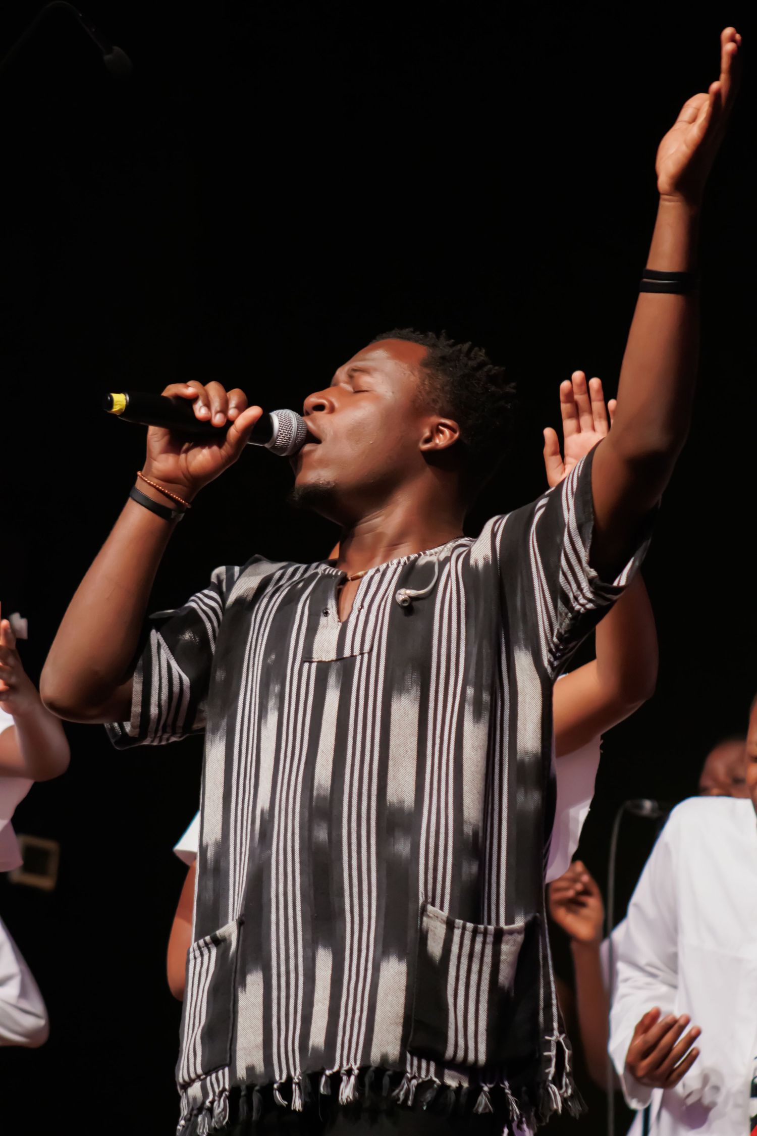 Performer in striped outfit singing with raised arms on dark stage.
