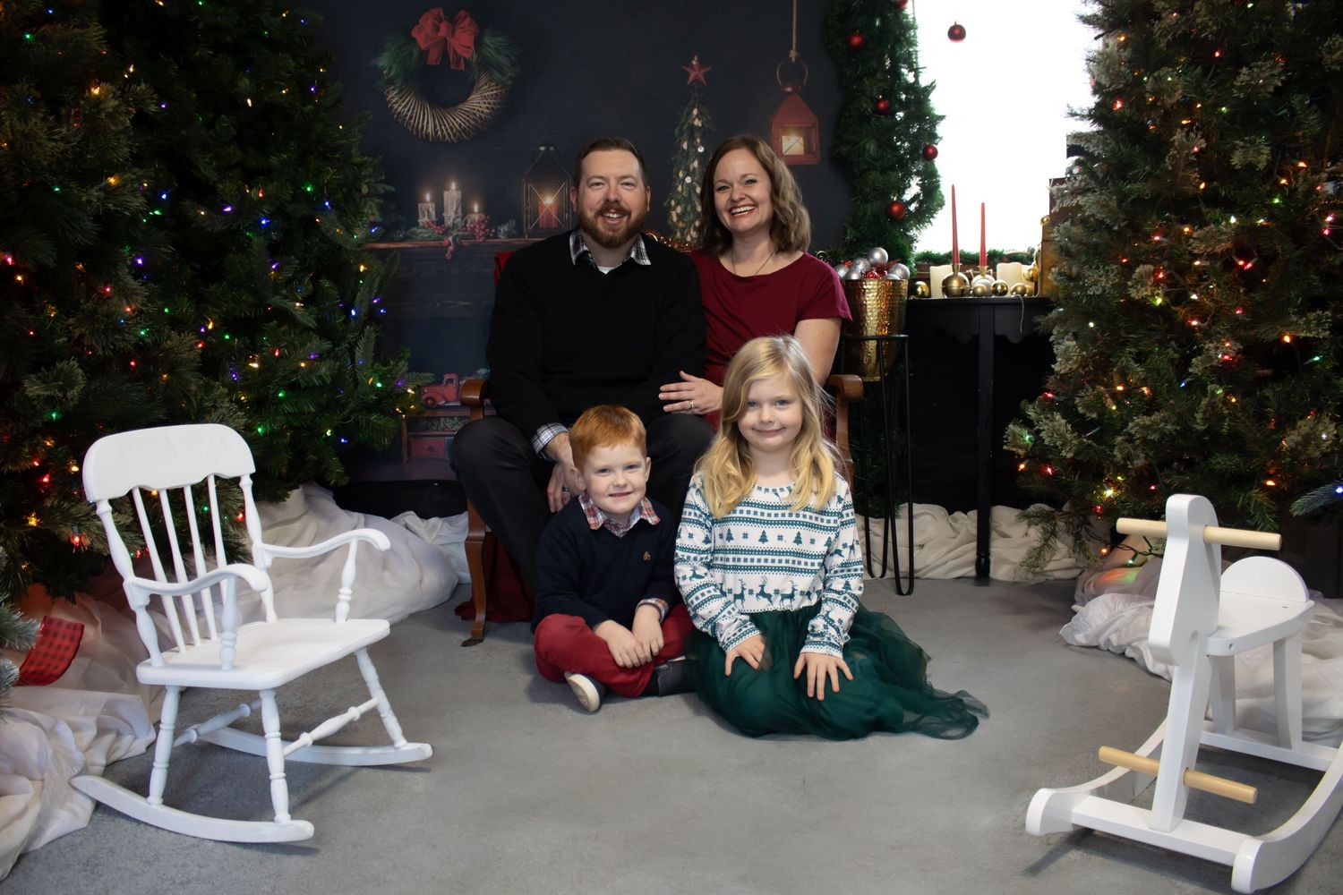 A family holiday portrait with Christmas trees and a white rocking chair in the background.