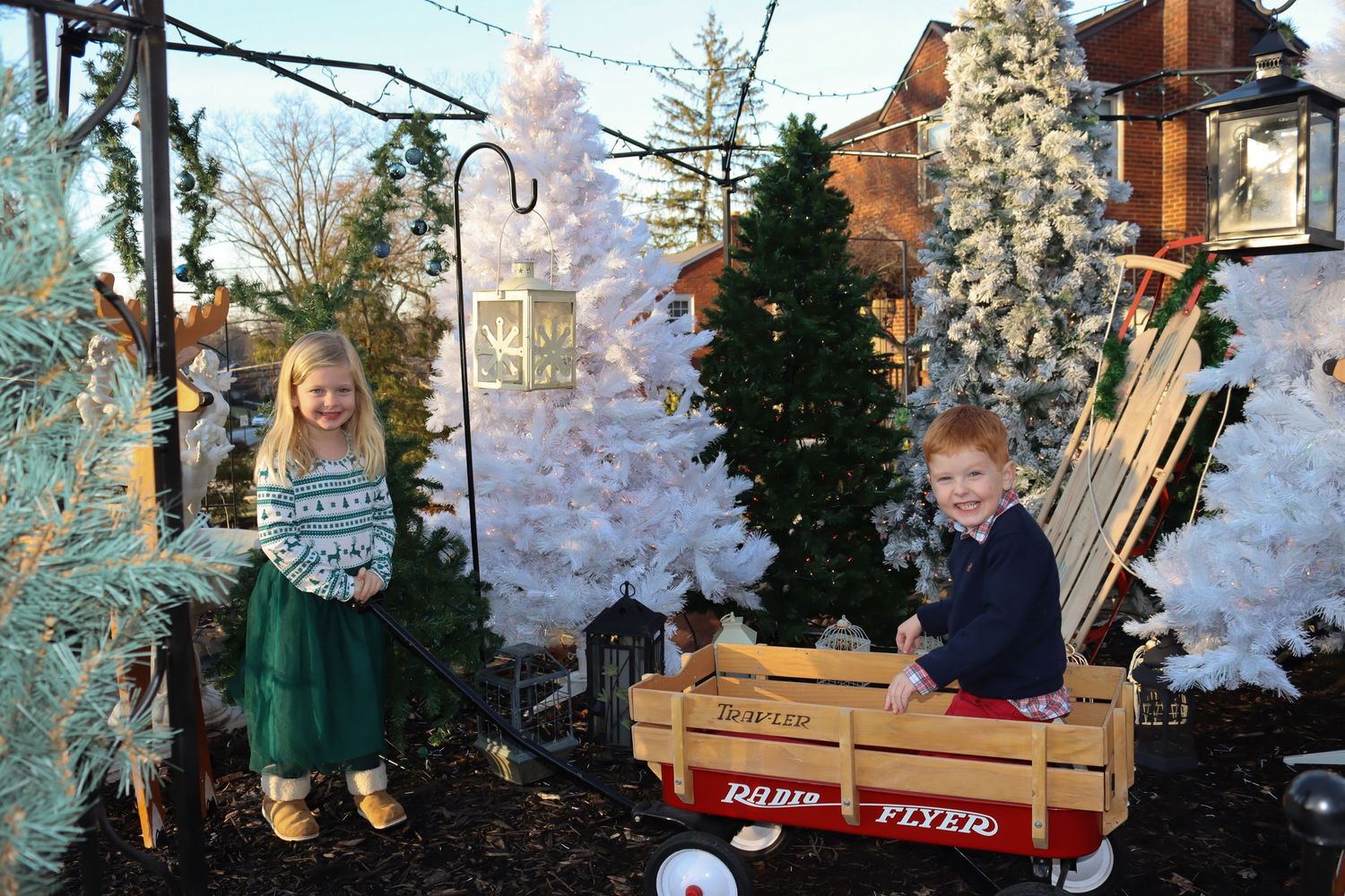 Two children pose with a red wagon among snow-flocked Christmas trees in an outdoor setting.