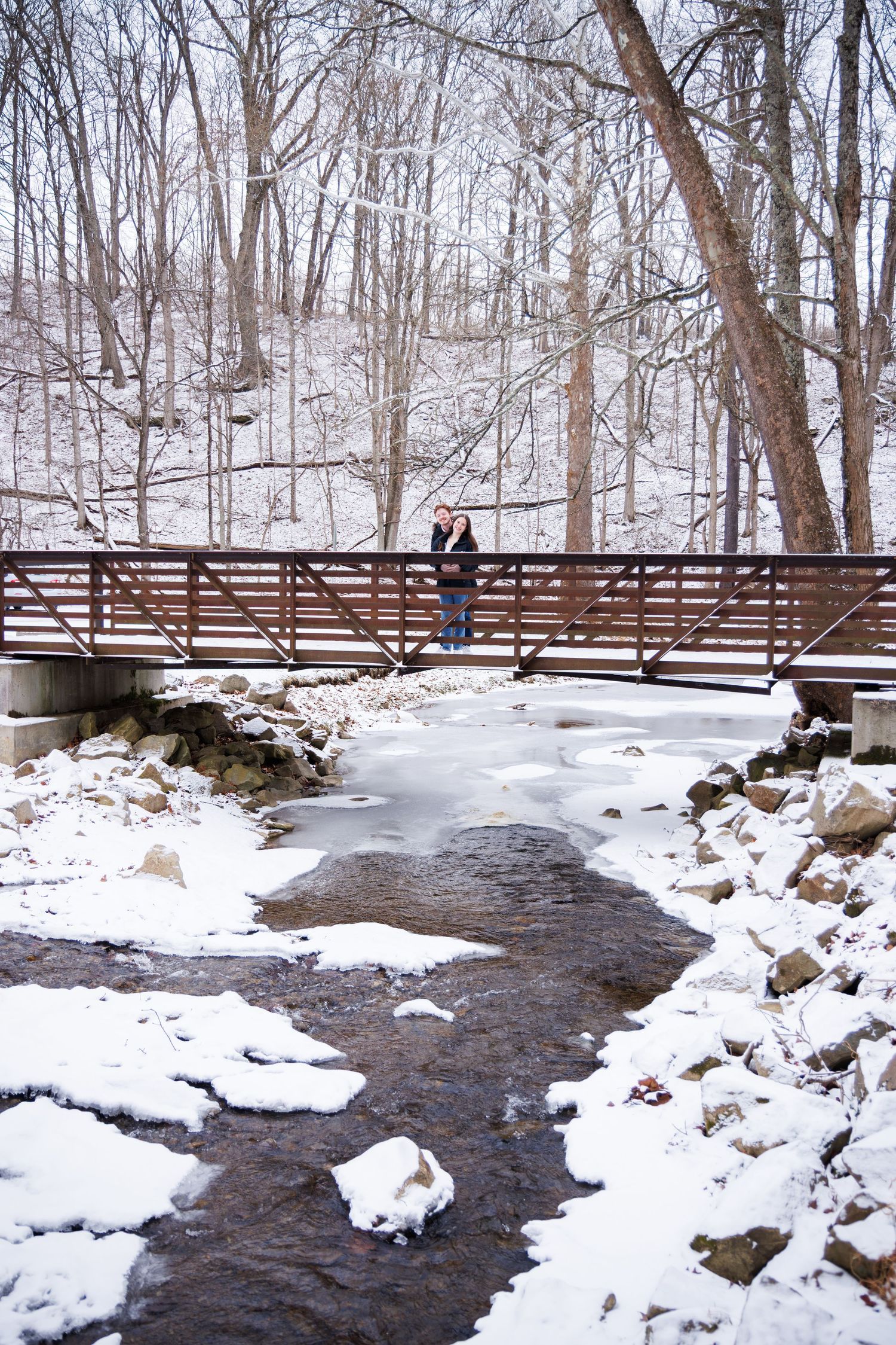A wooden footbridge spans across a flowing stream in a snow-covered winter forest landscape.