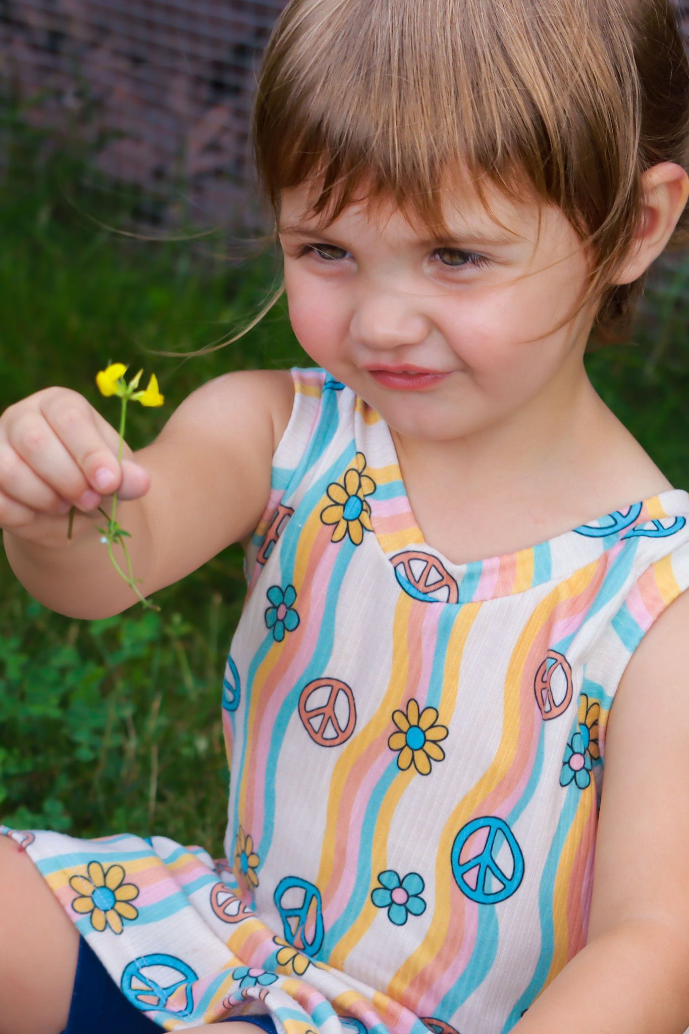 Child in striped peace sign dress holding small yellow flower in garden.