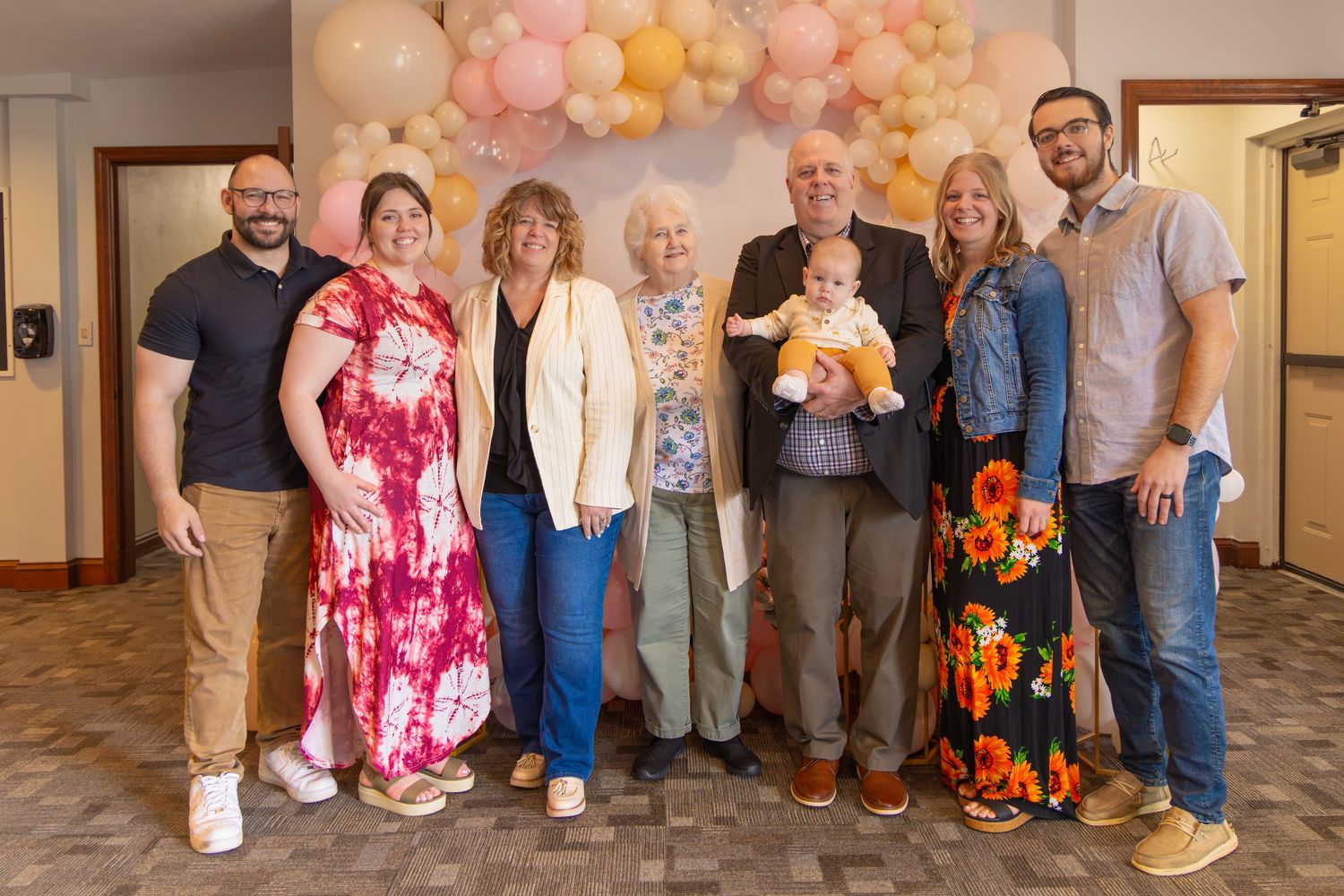 Family group photo with eight adults and a baby posing together at a celebration with balloon arch backdrop.