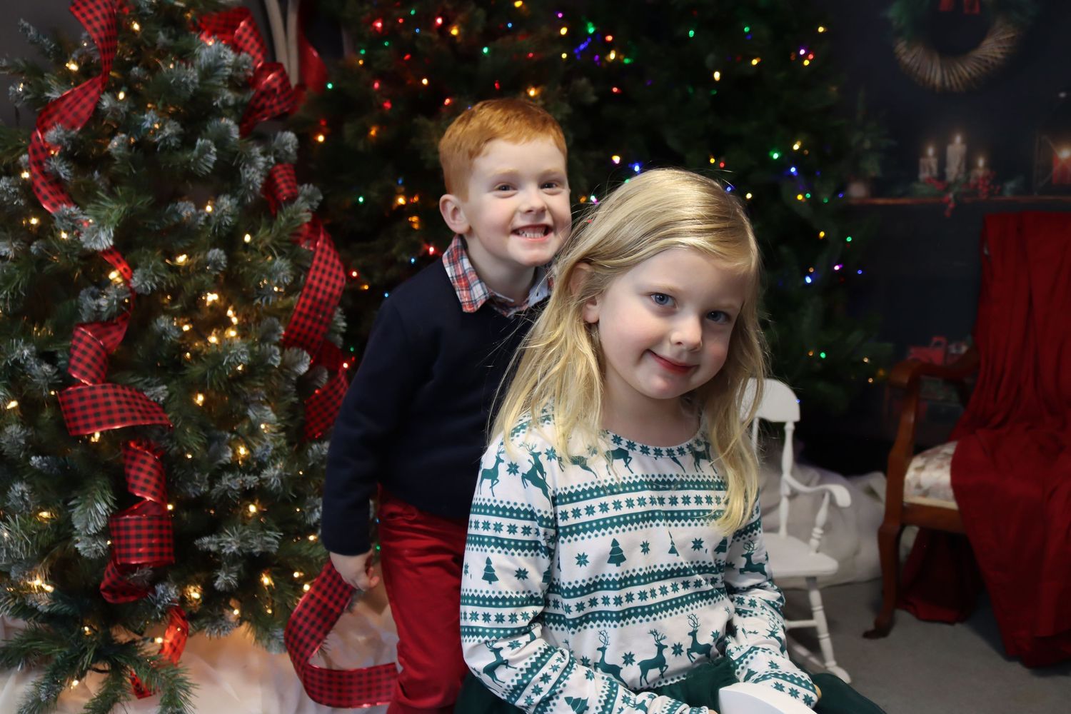 Two children pose in front of a decorated Christmas tree with twinkling lights and red ribbons.