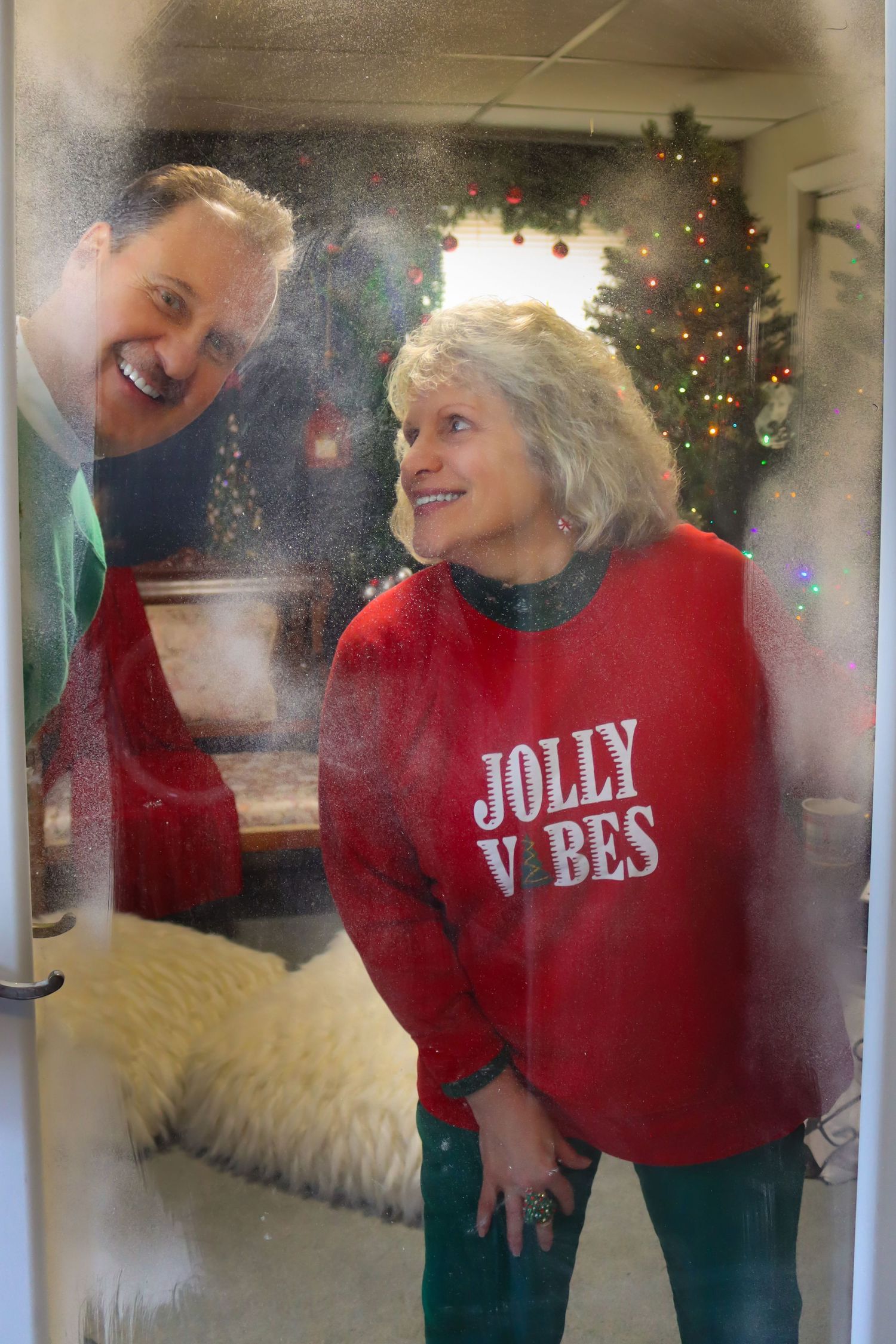 A cheerful holiday scene with two people wearing festive attire including a red 'Jolly Vibes' sweater near a Christmas tree.