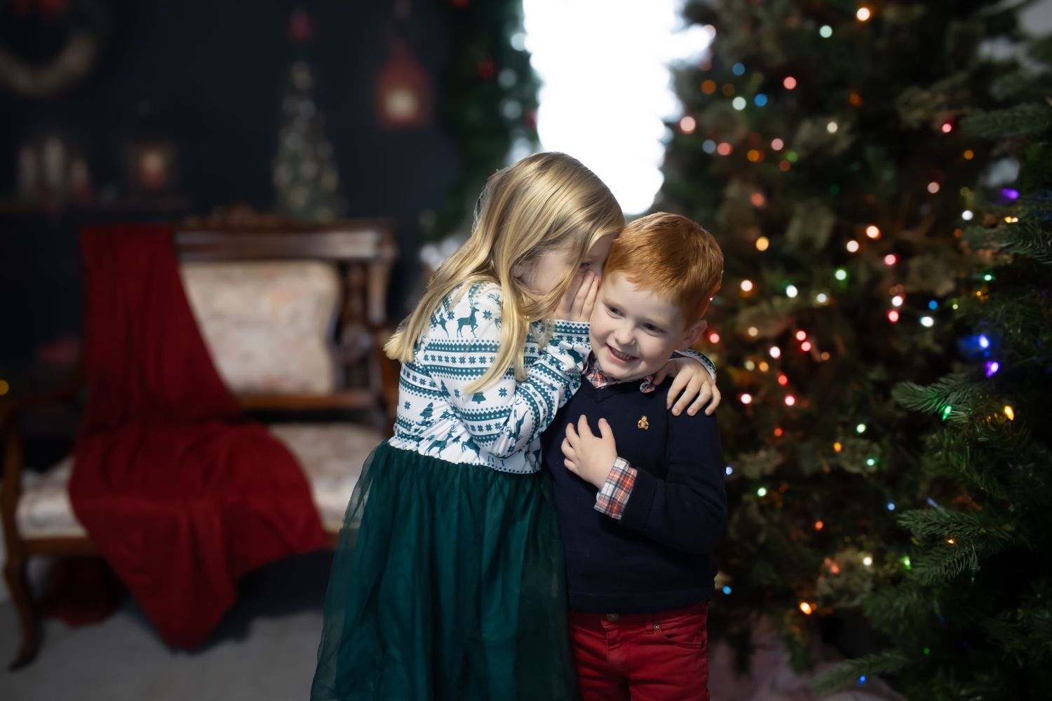 Two children share a warm embrace in front of a decorated Christmas tree.