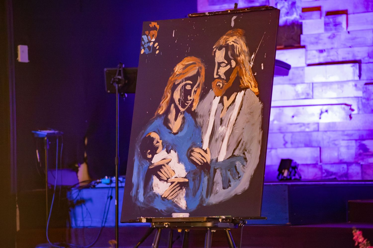 A painted artwork on an easel depicting the Holy Family with Mary, Joseph, and baby Jesus displayed at an event.