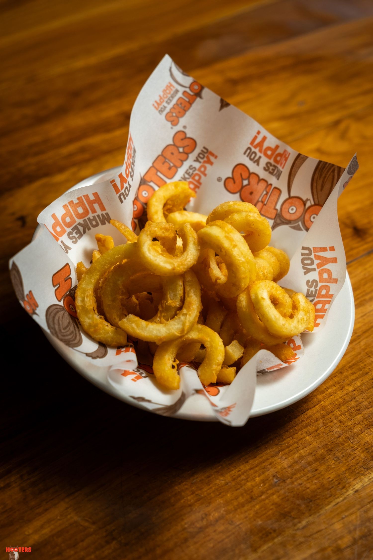 Golden curly fries served in branded paper on a white plate against a wooden table.