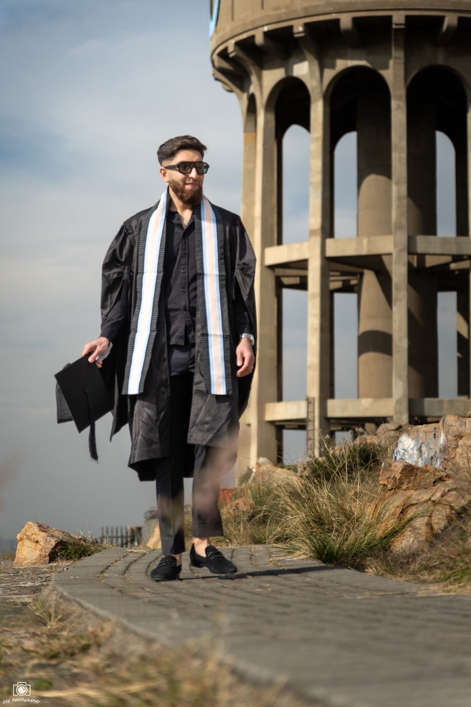 Graduate in cap and gown poses near architectural columns with city views.
