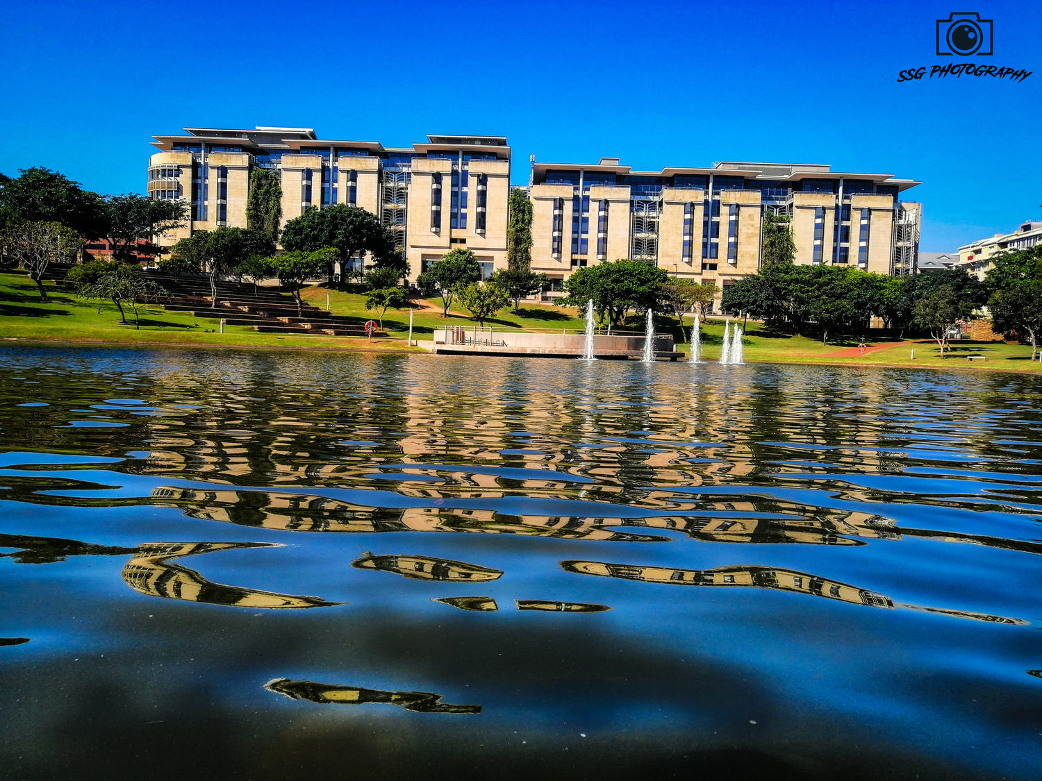 Modern apartment buildings reflect in rippling blue water with fountains at a lakeside residential complex.