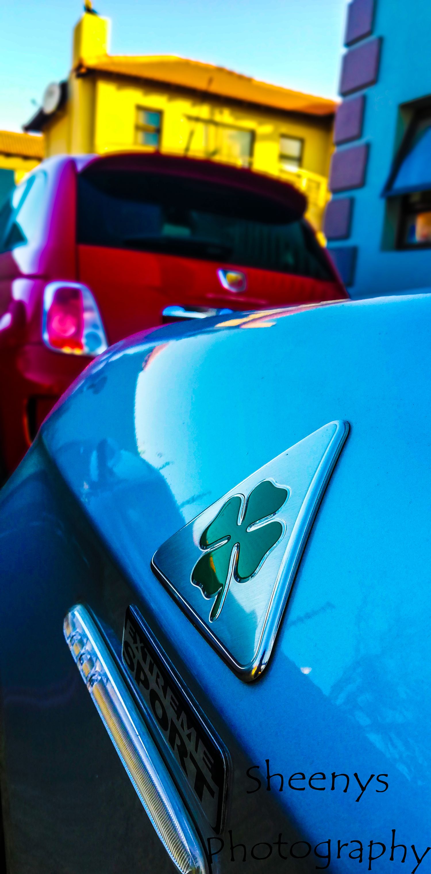Close-up of a blue Alfa Romeo with a four-leaf clover Quadrifoglio badge against colorful buildings in the background.