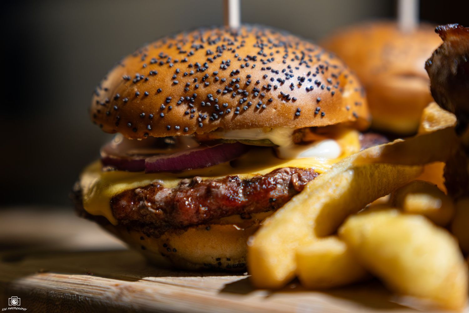 Close-up of a juicy cheeseburger with poppy seed bun and french fries on a rustic wooden surface.