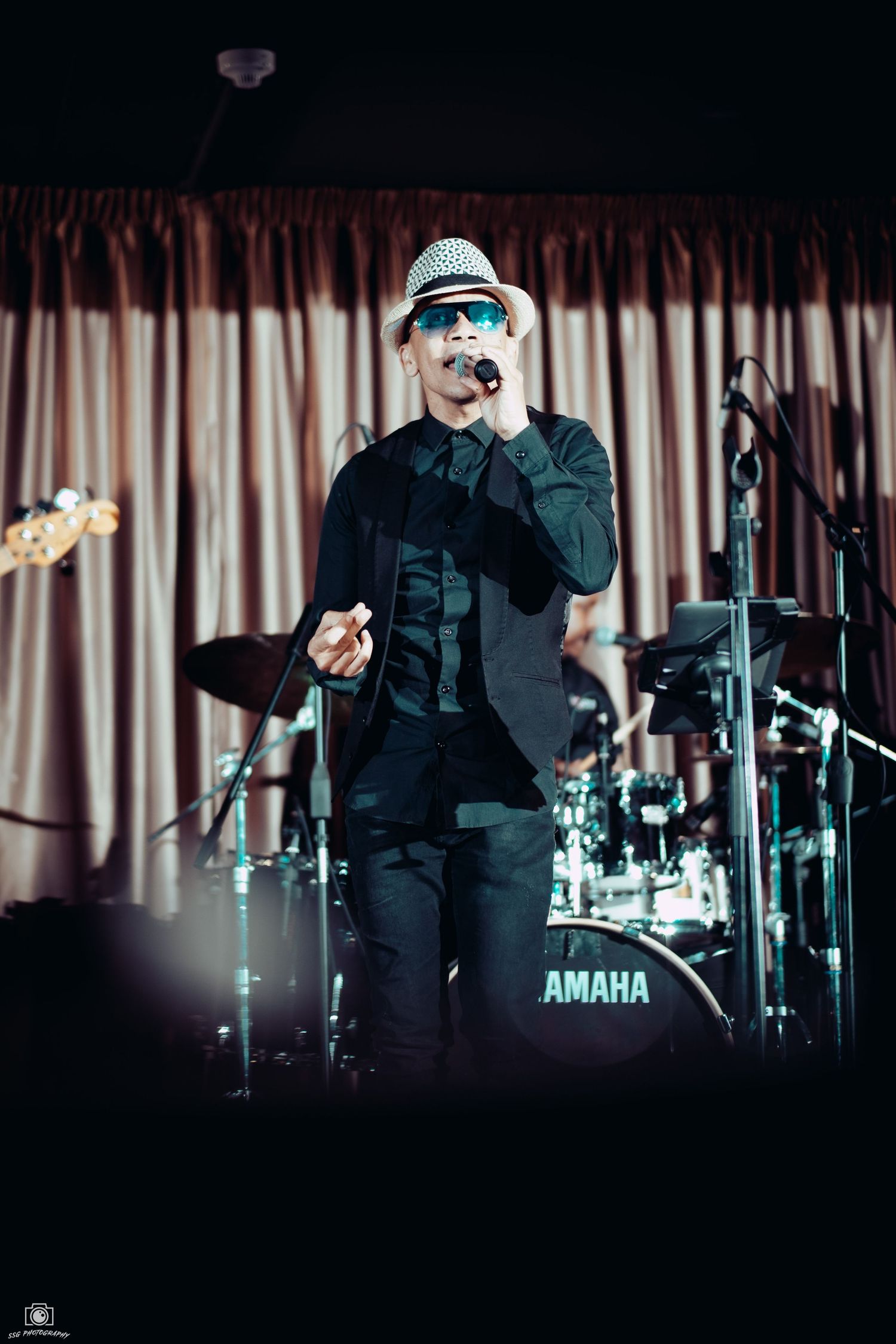 Singer in fedora and dark clothing performs in front of a Yamaha drum kit with curtained backdrop.